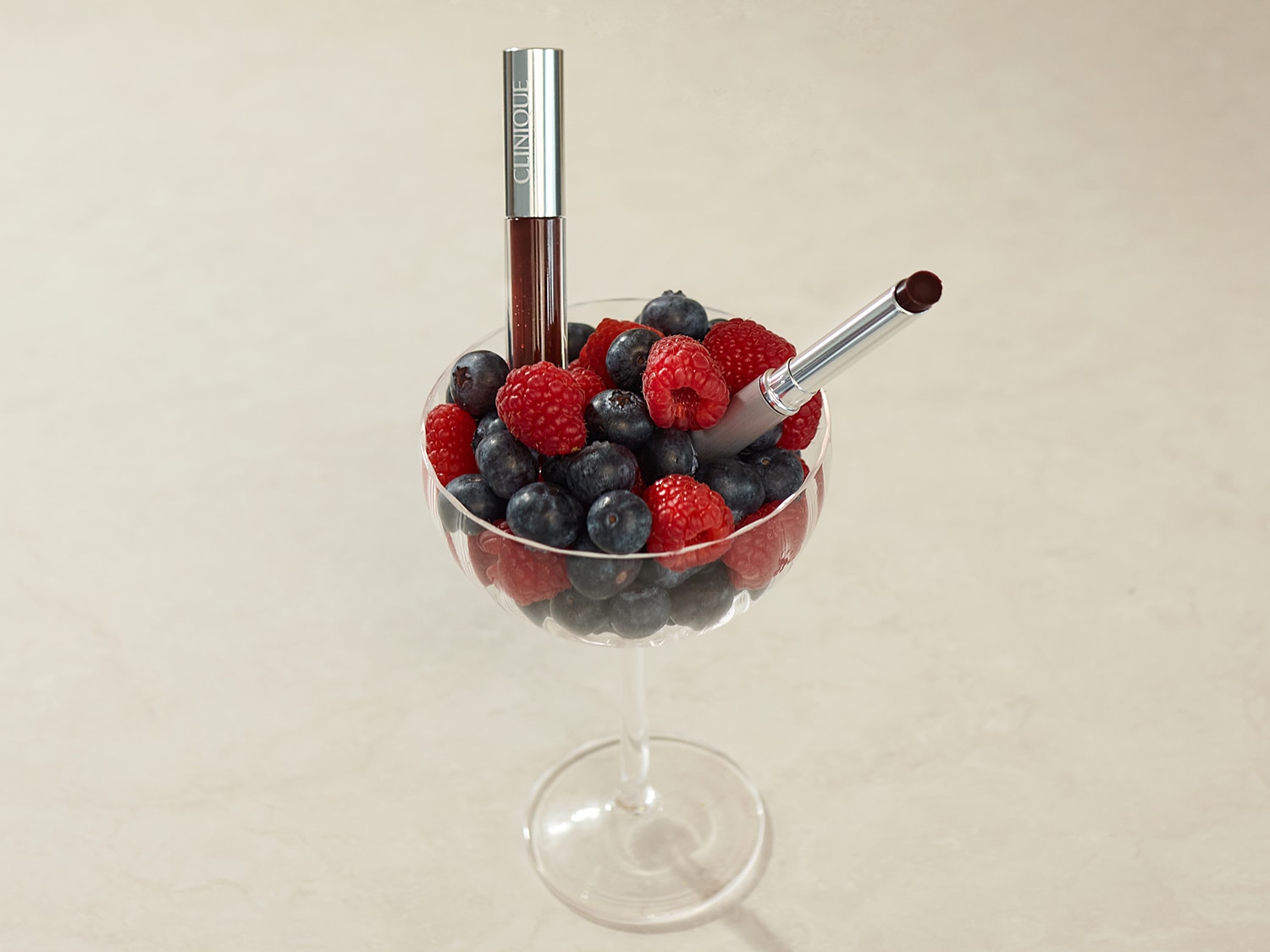 Een close-up van een wijnglas gevuld met bosbessen en frambozen, met een Clinique lippenstift en lipgloss in een zilveren verpakking.