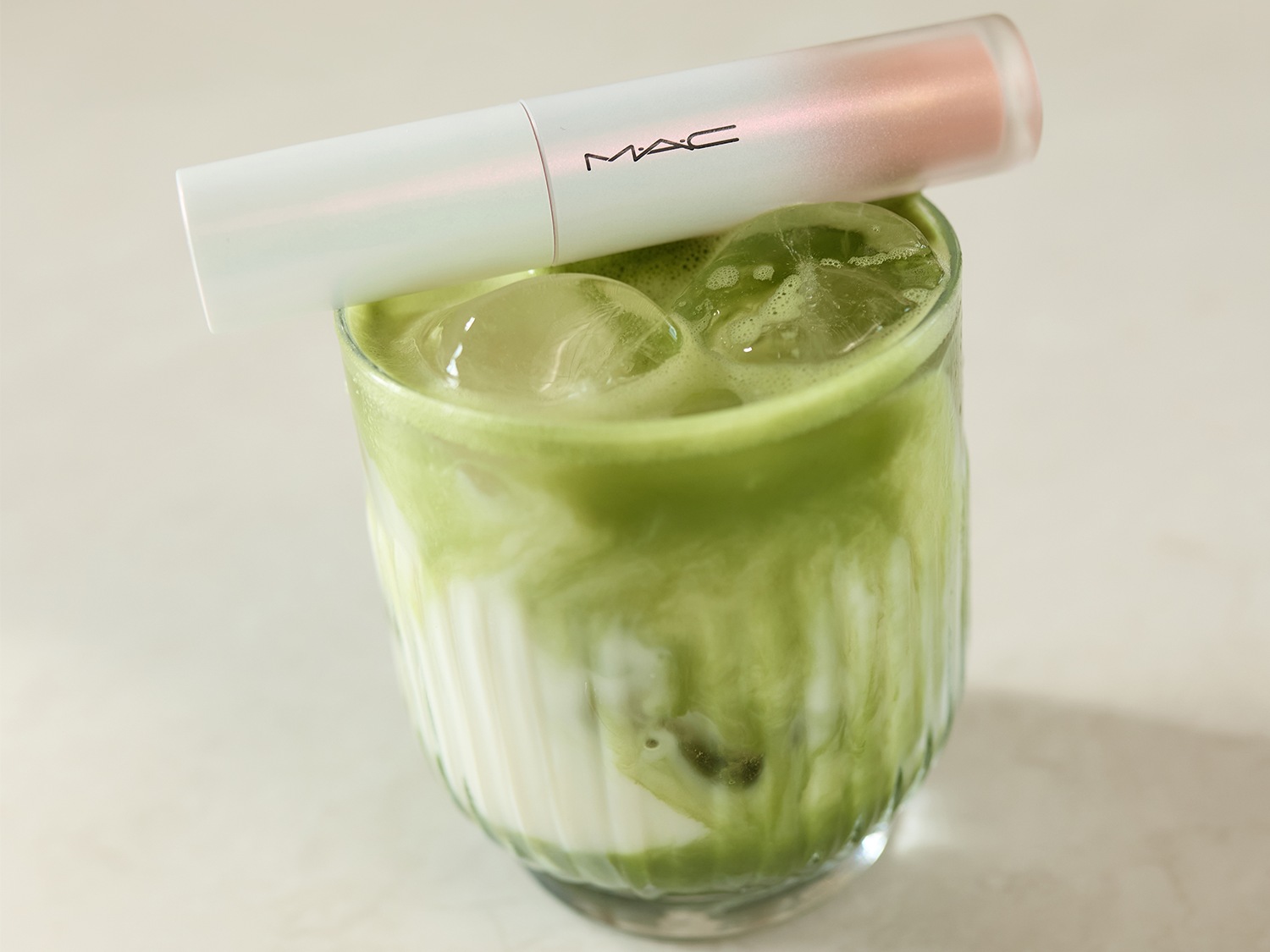 Een MAC lippenstift in een witte huls met een roze dop ligt bovenop een glas ijskoude matcha latte.