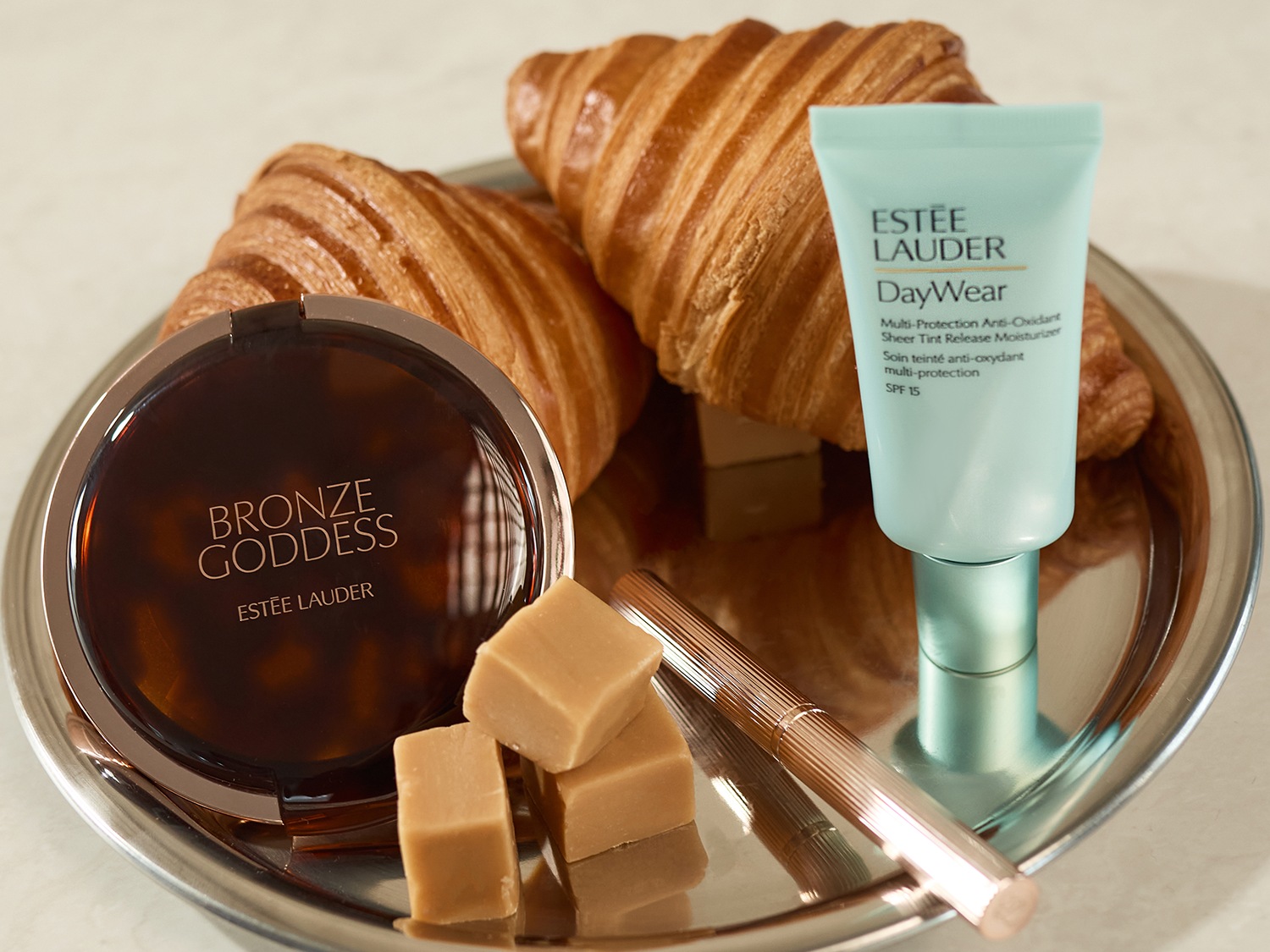 Op een zilveren schaal liggen croissants, karamelblokjes en Estée Lauder-producten, waaronder Bronze Goddess en DayWear Multi-Protection Anti-Oxidant Sheer Tint Release Moisturizer SPF 15.