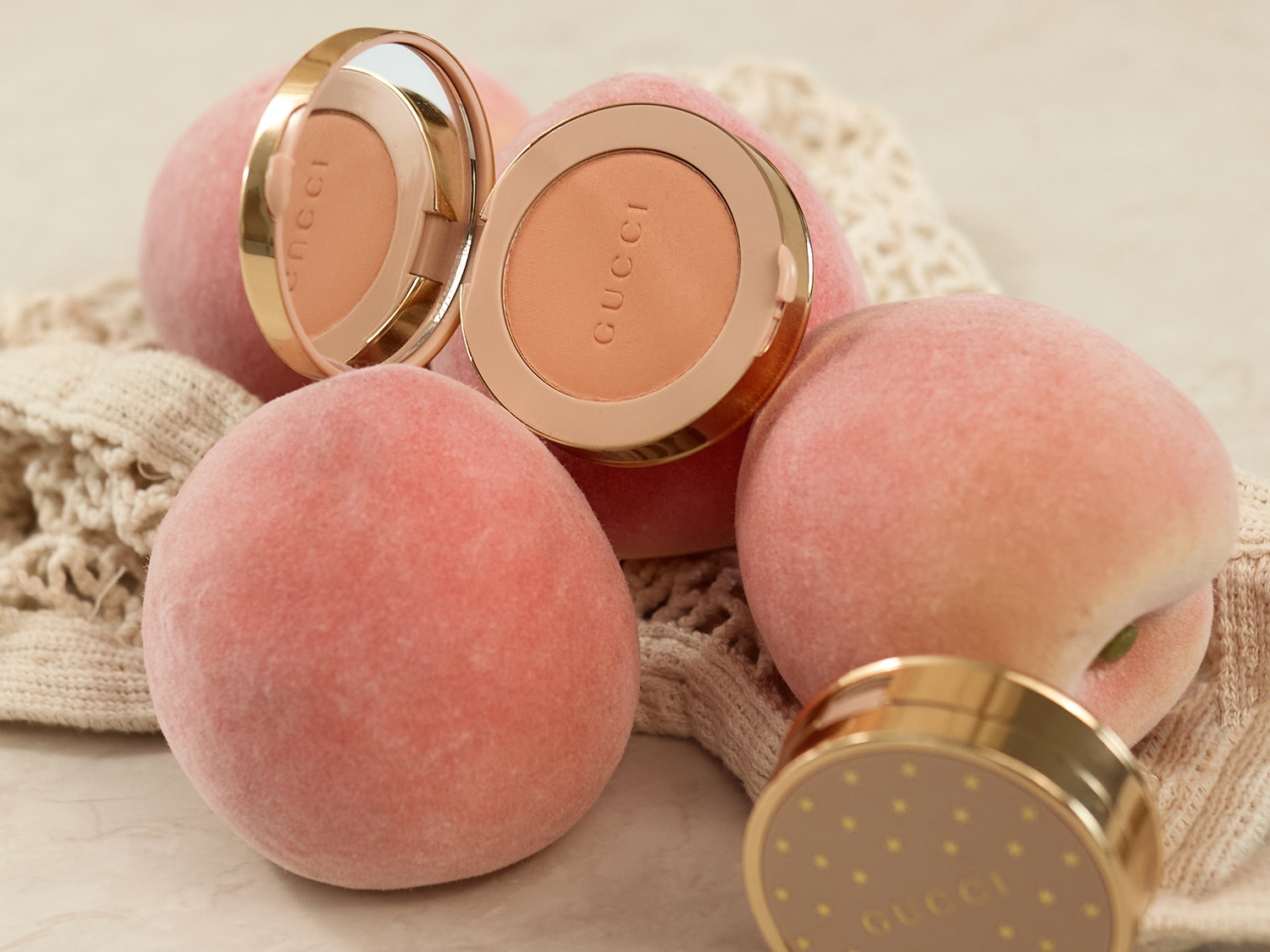 Gucci compact poeder met gouden rand en perzikkleurige tint, geplaatst tussen zachte perziken en een gebreide doek.