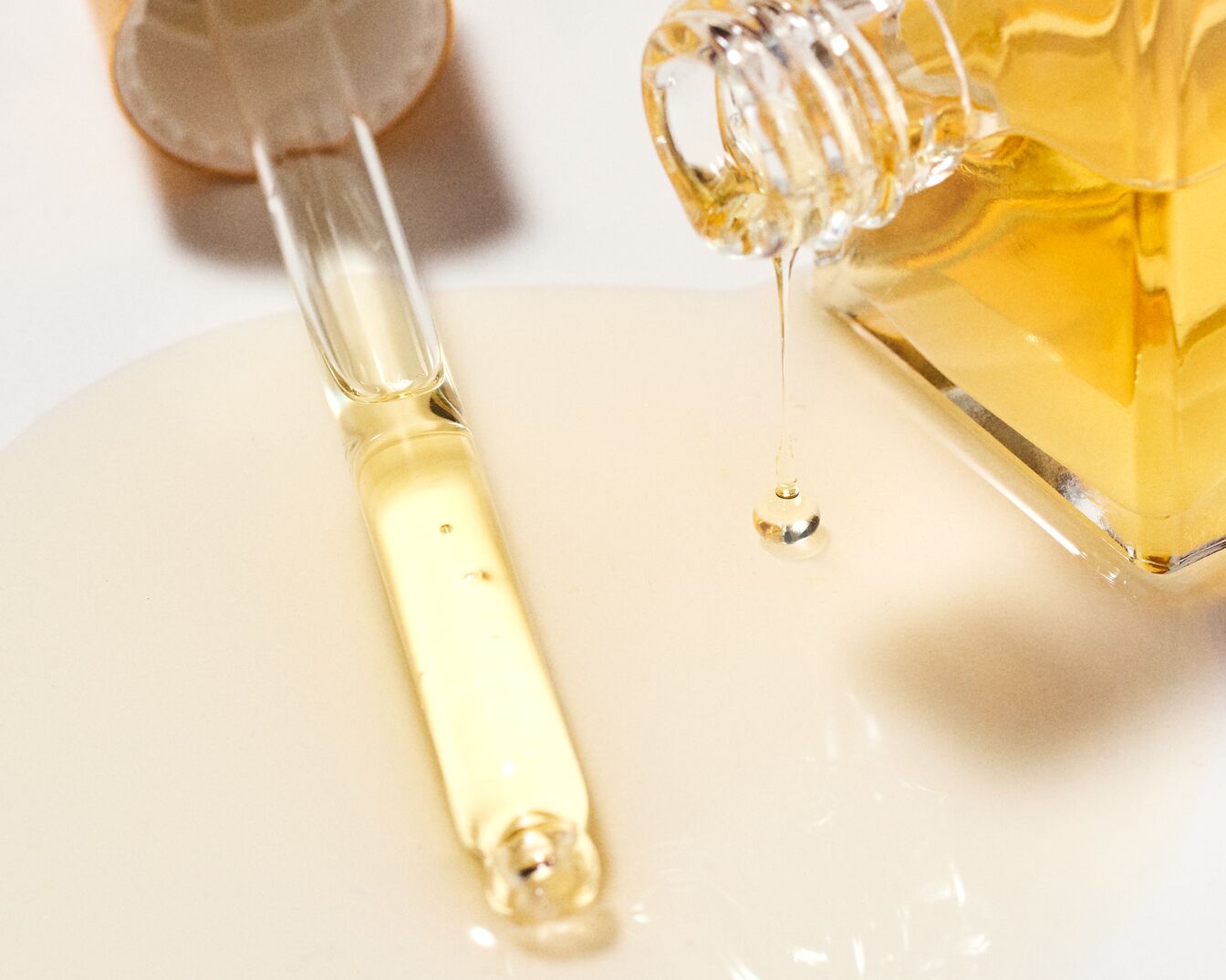 Nahaufnahme eines klaren Glasfläschchens mit goldfarbenem Öl, das ausläuft, daneben eine Pipette, die ebenfalls mit dem Öl gefüllt ist, auf einem hellen Untergrund.