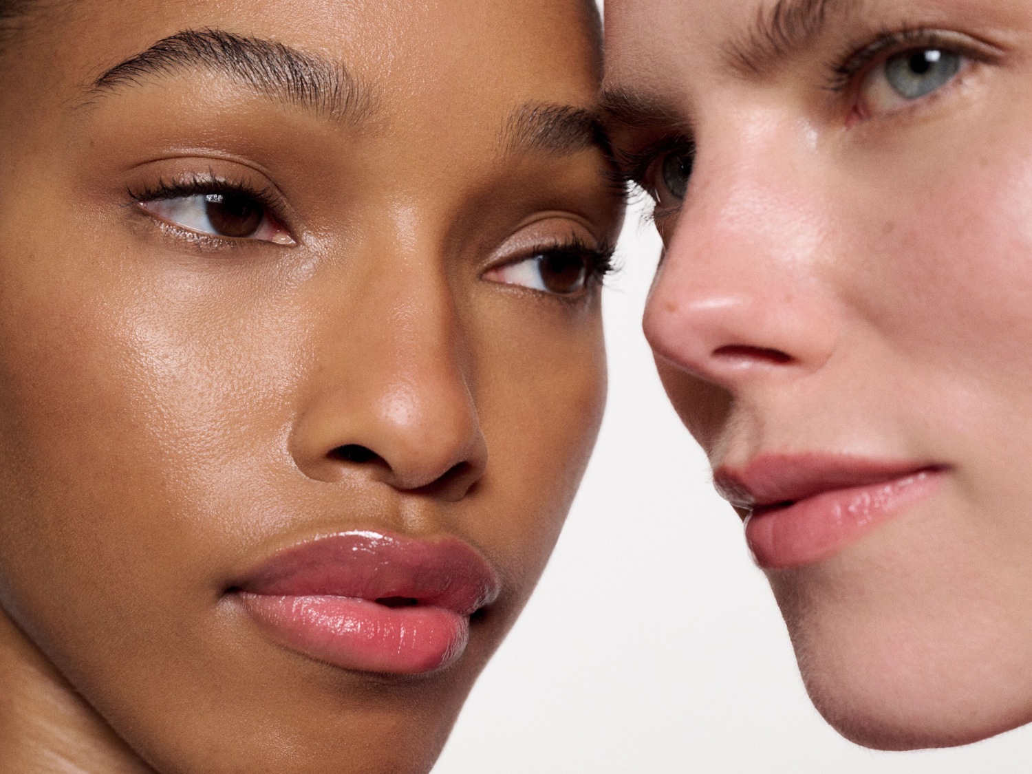 Primo piano di due donne con pelle luminosa e labbra lucide, una con la pelle scura e l'altra chiara, che mostra la diversità e la bellezza naturale.