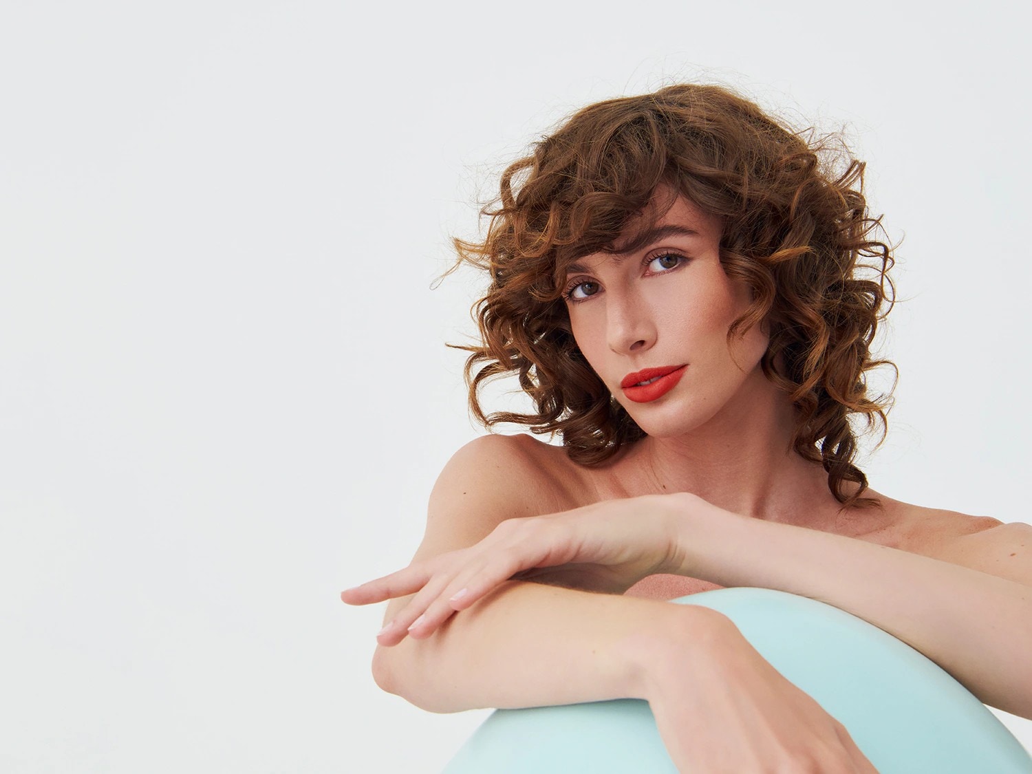 Porträt einer Frau mit lockigem, braunem Haar und roten Lippen, die auf einem hellblauen Stuhl sitzt und in die Kamera schaut.