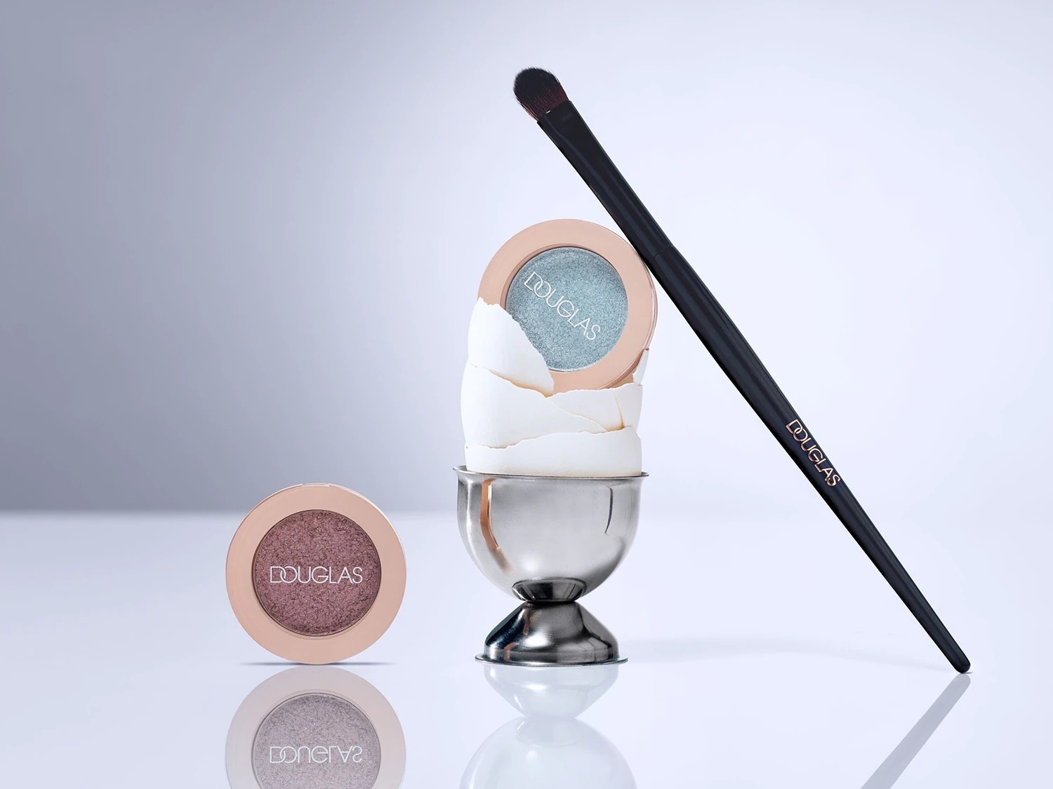 Douglas Lidschatten in Rosé und Türkis, präsentiert mit einem schwarzen Pinsel und einem zerbrochenen Ei in einem silbernen Eierbecher.