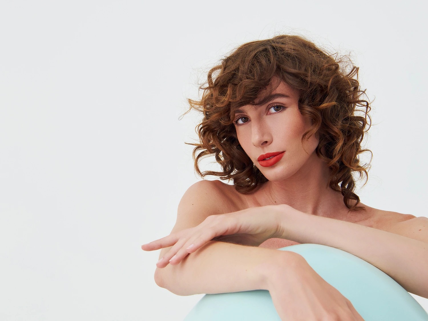 Porträt einer Frau mit lockigem, braunem Haar und roten Lippen, die auf einem hellblauen Gegenstand sitzt und in die Kamera blickt.