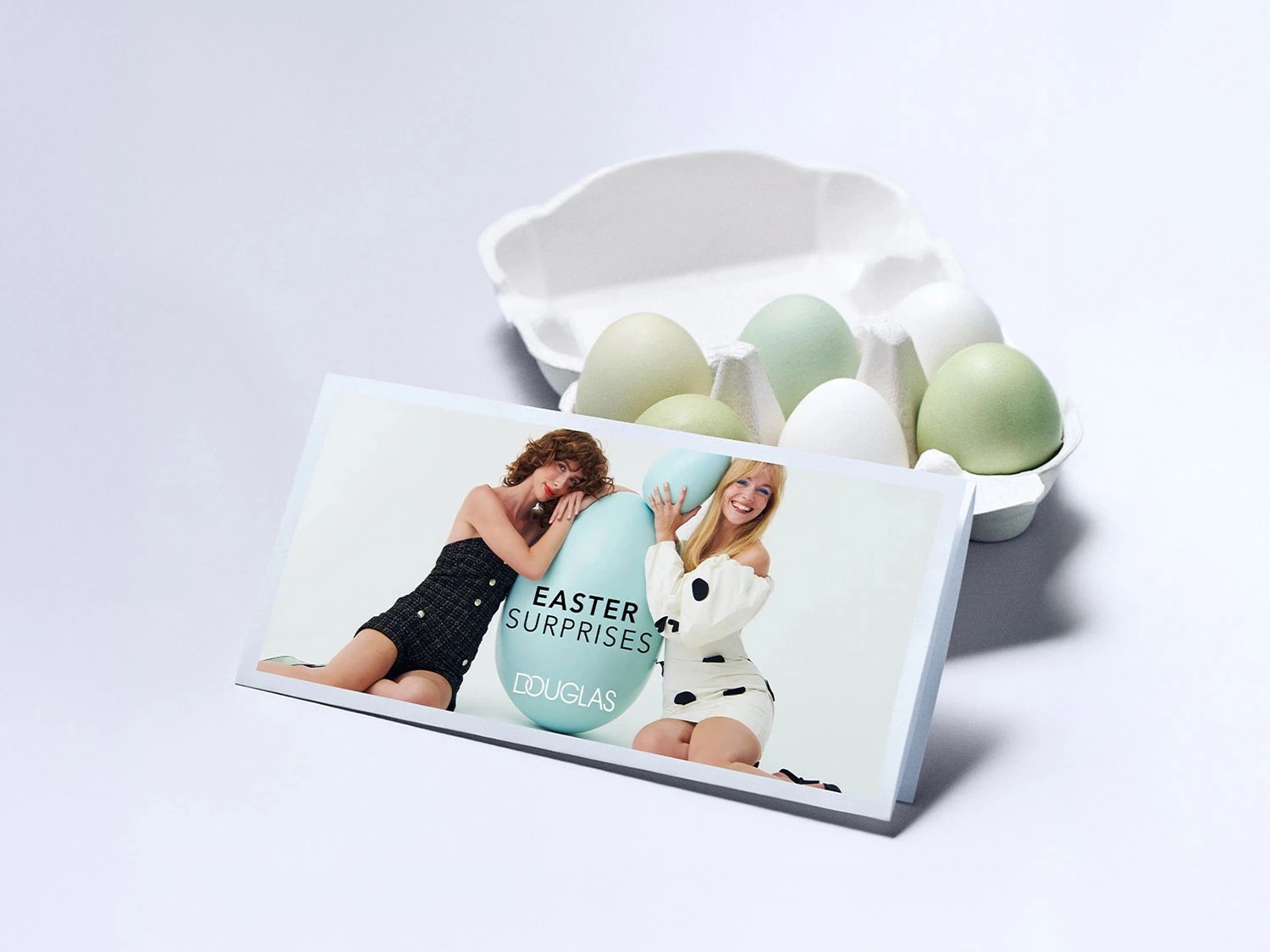 Ostereiersuche mit Douglas: Zwei Frauen posieren mit pastellfarbenen Ostereiern vor einem Hintergrund aus echten Eiern in einem Eierkarton, die 'Easter Surprises' versprechen.