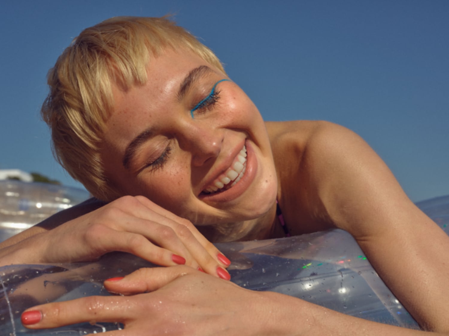 Una donna sorridente con i capelli biondi corti e un tratto di eyeliner blu brillante, rilassata su un galleggiante trasparente sotto il sole.