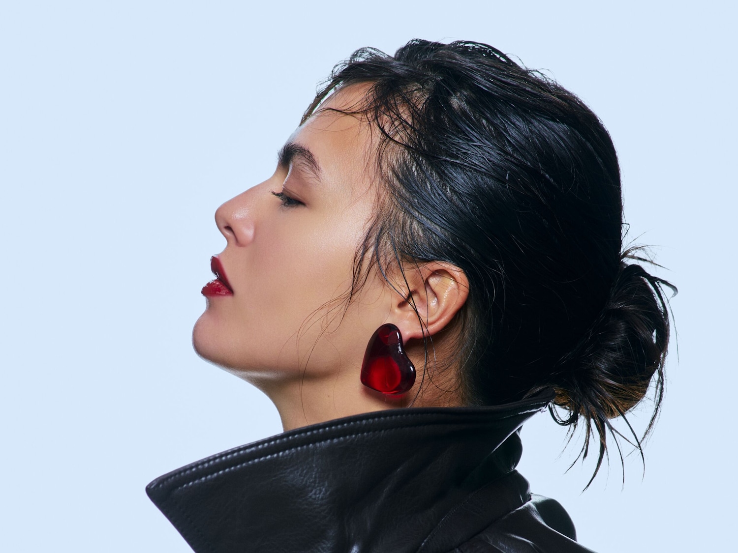 Una donna con i capelli scuri raccolti, labbra rosse lucide e orecchini a forma di cuore rosso, indossa una giacca di pelle nera su uno sfondo azzurro chiaro.