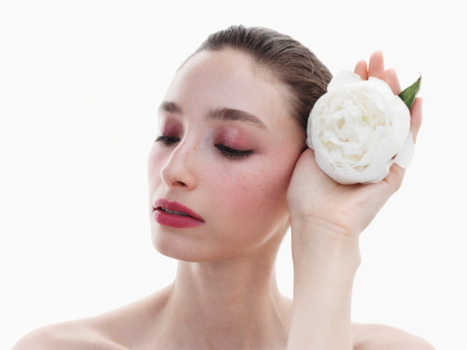 Primo piano di una donna con ombretto rosa e labbra rosse, che tiene un fiore bianco vicino all'orecchio, incarnando la bellezza e la delicatezza.