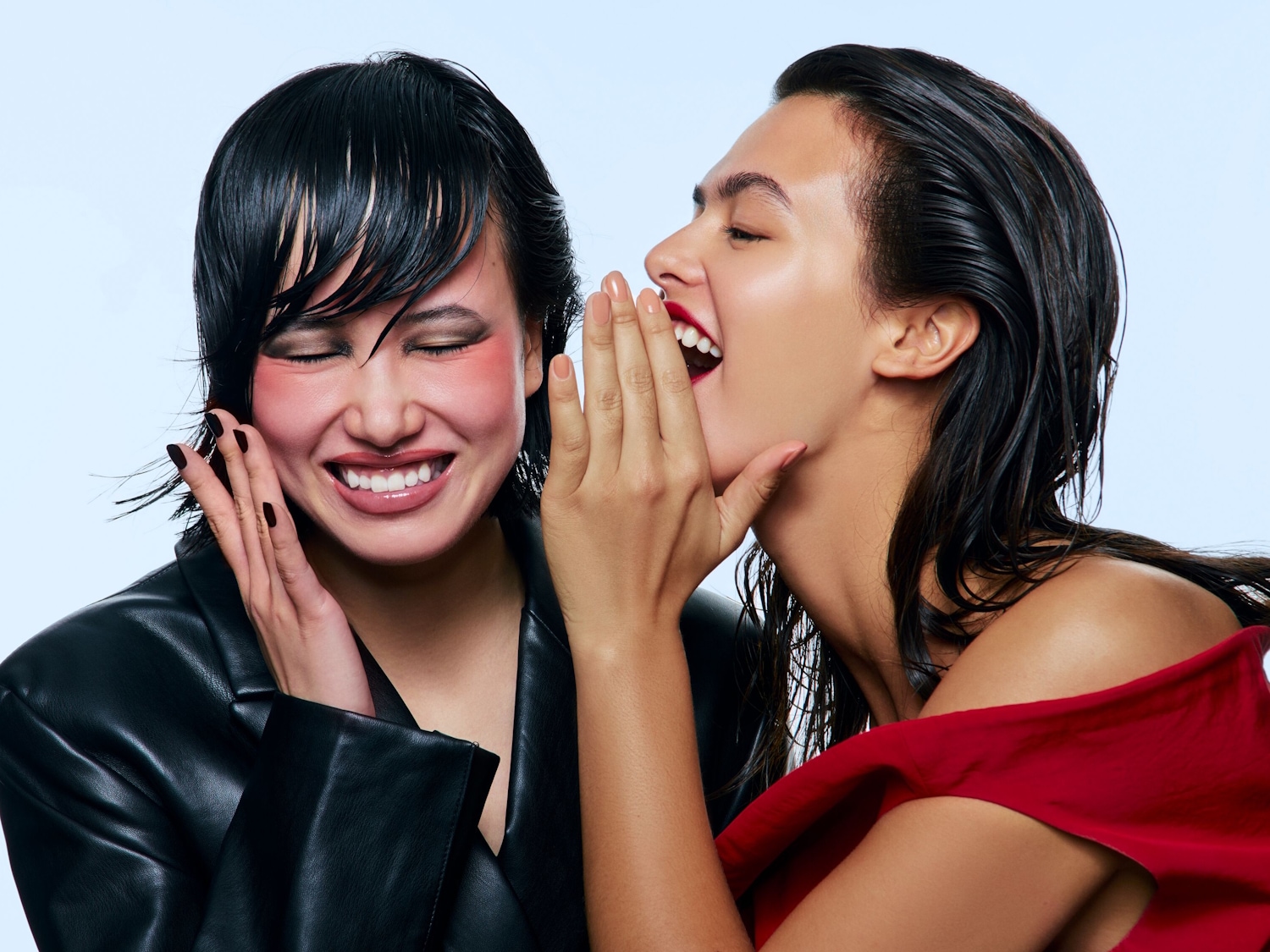 Due donne ridono, una con un blazer nero e fard rosa acceso, l'altra con un top rosso e labbra scarlatte, mentre si sussurrano un segreto.