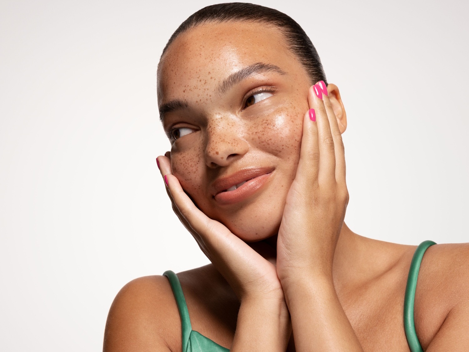 Primo piano di una donna con lentiggini, pelle luminosa e un top verde, che appoggia le mani sul viso e guarda di lato.