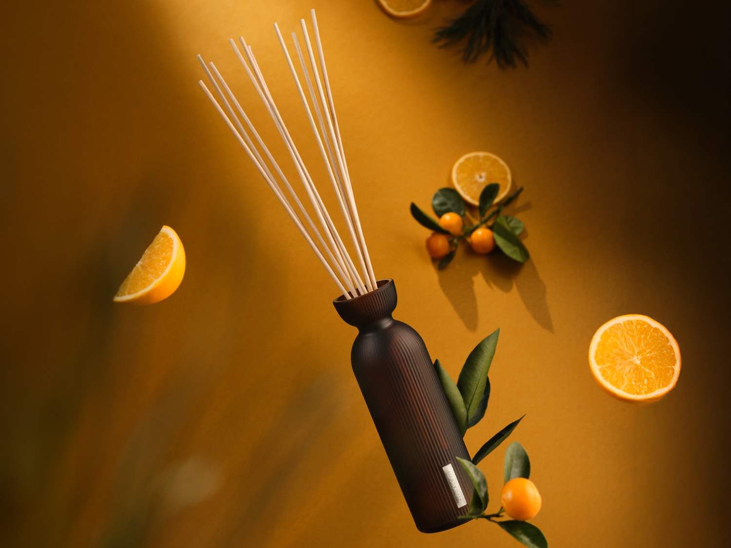 Un diffuseur de parfum marron foncé avec des bâtonnets est entouré de tranches d'orange et de feuilles vertes sur un fond jaune.