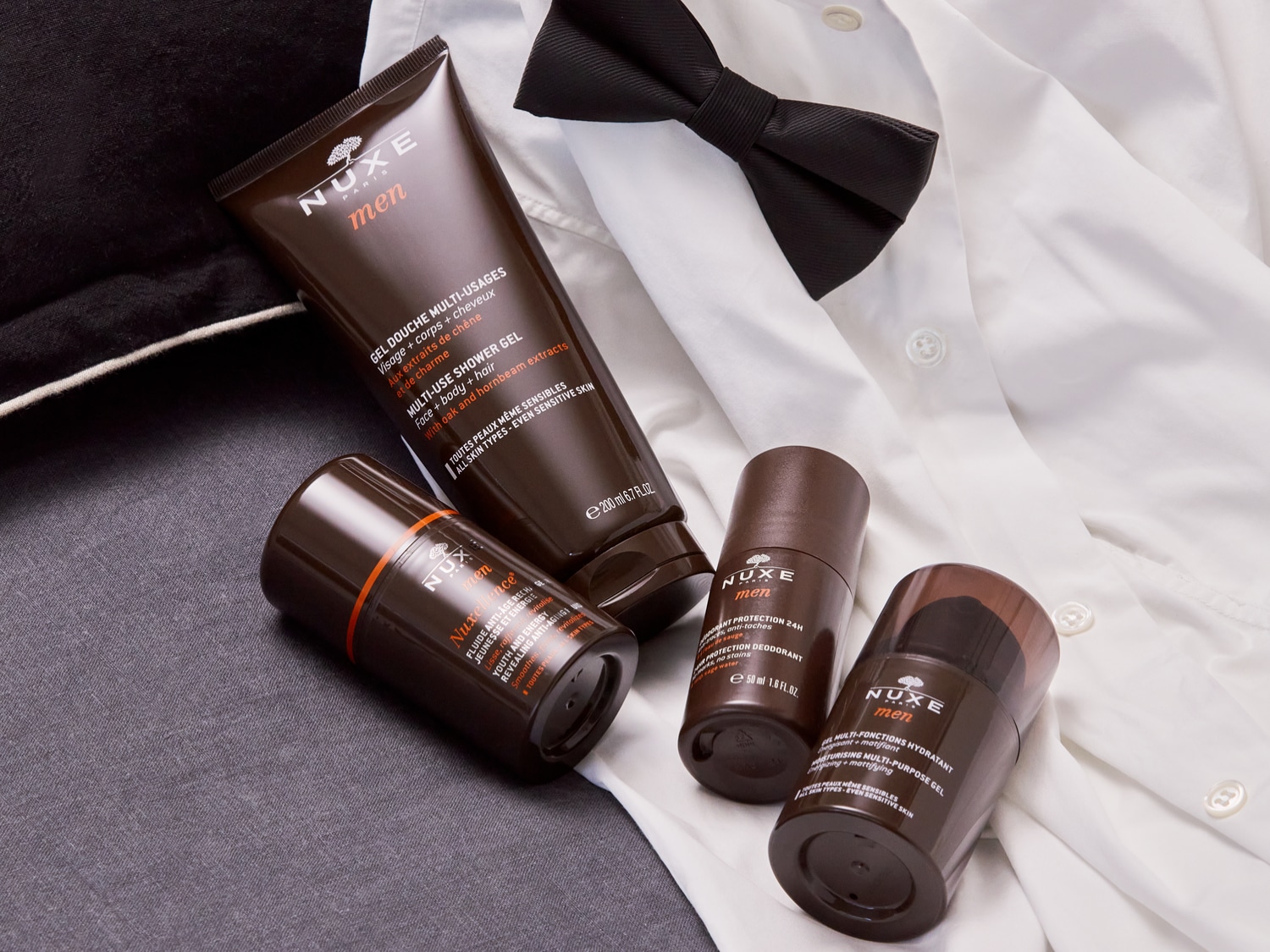 Nuxe Men-collectie met multi-use showergel, anti-aging fluïde, 24-uurs deodorant en hydraterende multi-purpose gel, gepresenteerd op een witte blouse met een zwarte vlinderdas.