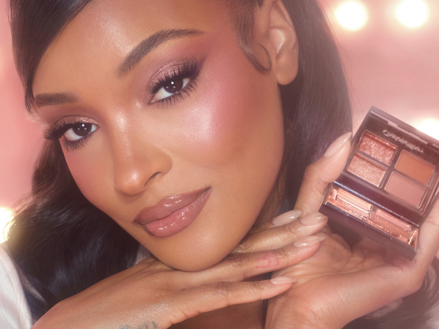 Nahaufnahme einer Frau mit schimmerndem rosa Lidschatten und glänzenden Lippen, die eine kleine Lidschattenpalette in der Hand hält, umgeben von einem weichen, rosa Hintergrund.