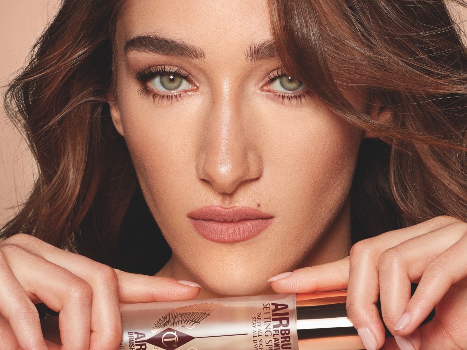 Nahaufnahme einer Frau mit hellbraunem, gewelltem Haar und grünen Augen, die Charlotte Tilbury Airbrush Flawless Setting Spray in den Händen hält.