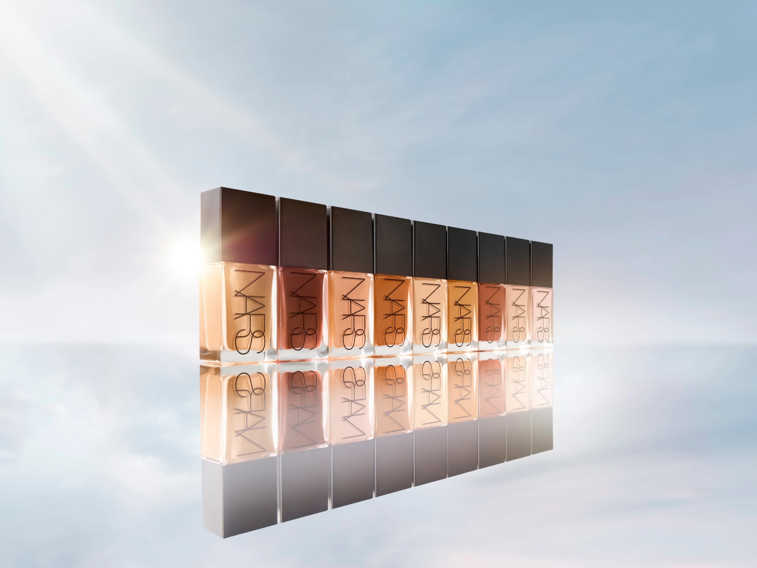 Een rij NARS foundationflessen in verschillende tinten, van licht naar donker, met zwarte doppen en een heldere, reflecterende basis.