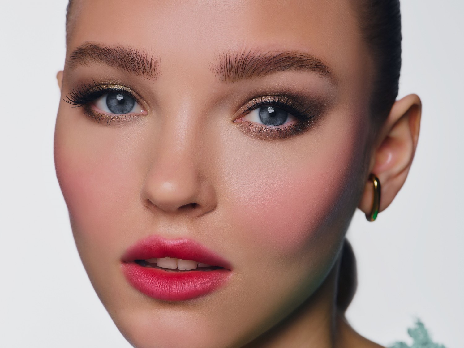 Una donna con un trucco audace, che presenta un ombretto dorato scintillante, un eyeliner scuro, ciglia voluminose, sopracciglia definite, un blush rosa sulle guance e labbra rosse intense, con un orecchino a cerchio dorato visibile sull'orecchio destro.