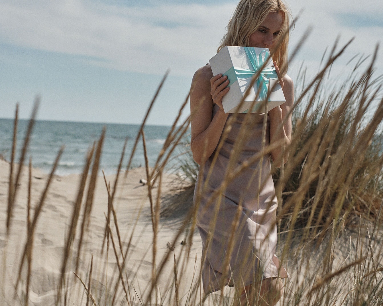 Eine blonde Frau in einem hellbraunen Kleid steht am Strand und hält eine weiße Geschenkbox mit einer hellblauen Schleife vor ihr Gesicht.