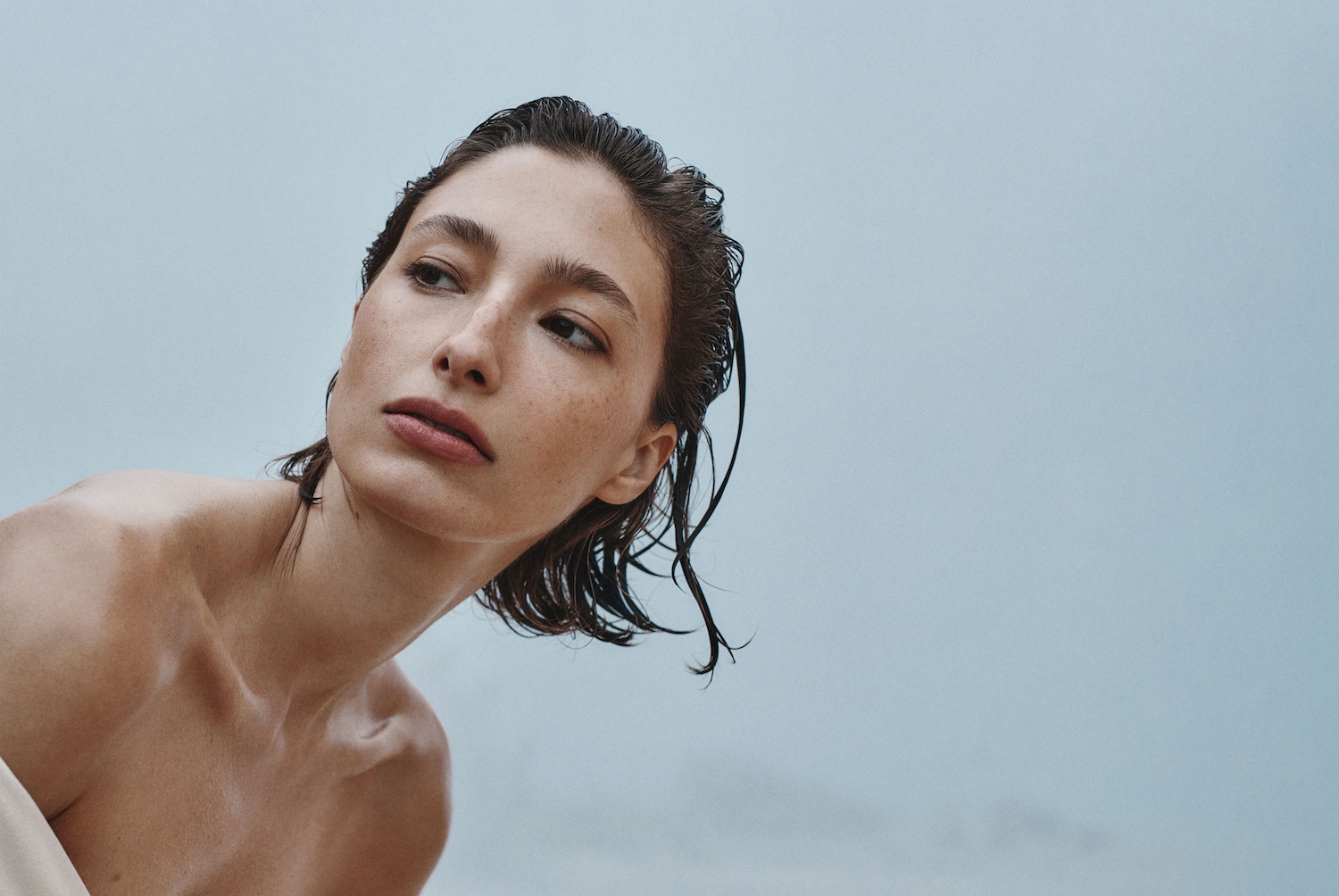 Porträt einer jungen Frau mit nassen, dunklen Haaren und Sommersprossen, die über ihre Schulter in Richtung des hellblauen Hintergrunds blickt.