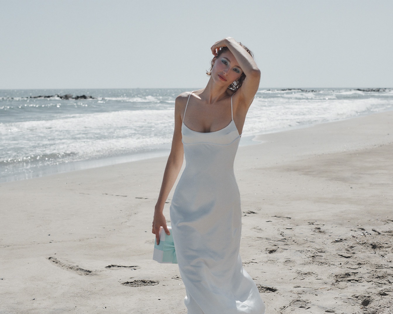 Eine Frau im weißen Kleid steht am Strand und hält eine hellblaue Schachtel in der Hand, während die Wellen sanft an den Sand rollen.