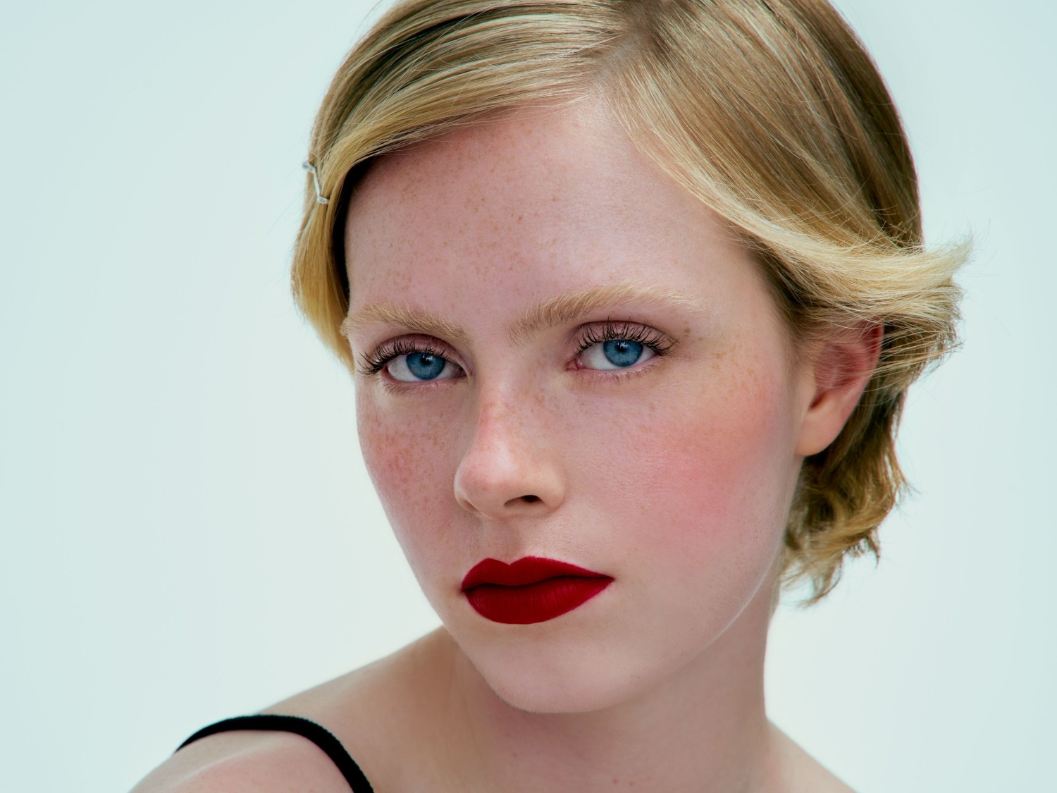 Nahaufnahme eines blassen, sommersprossigen Gesichts einer Frau mit roten Lippen, blonden Haaren, hellen Augenbrauen und blauen Augen.