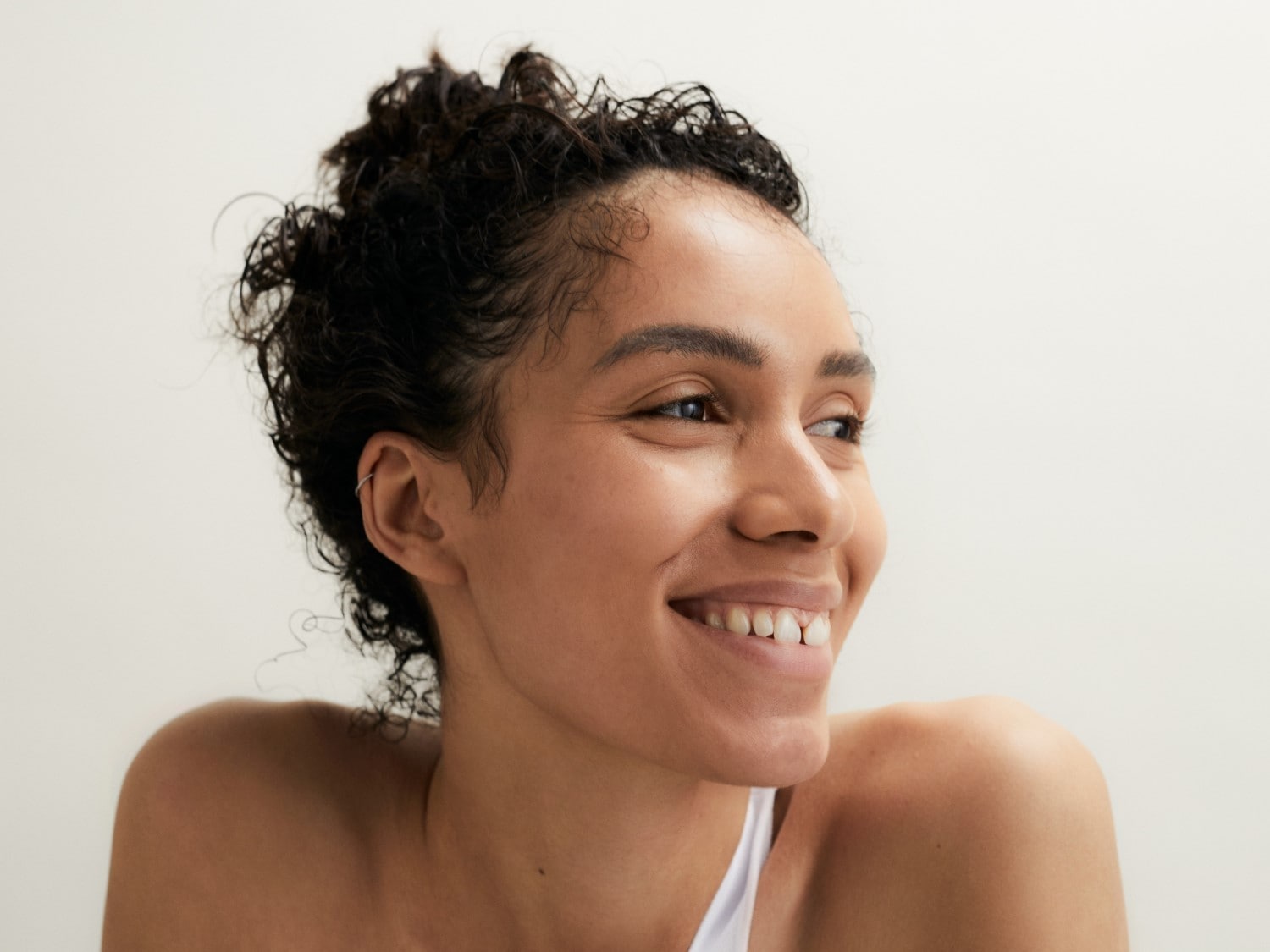 Nahaufnahme einer strahlenden Frau mit lockigem, dunklem Haar, die lächelt und zur Seite blickt, während sie ein weißes Top trägt.