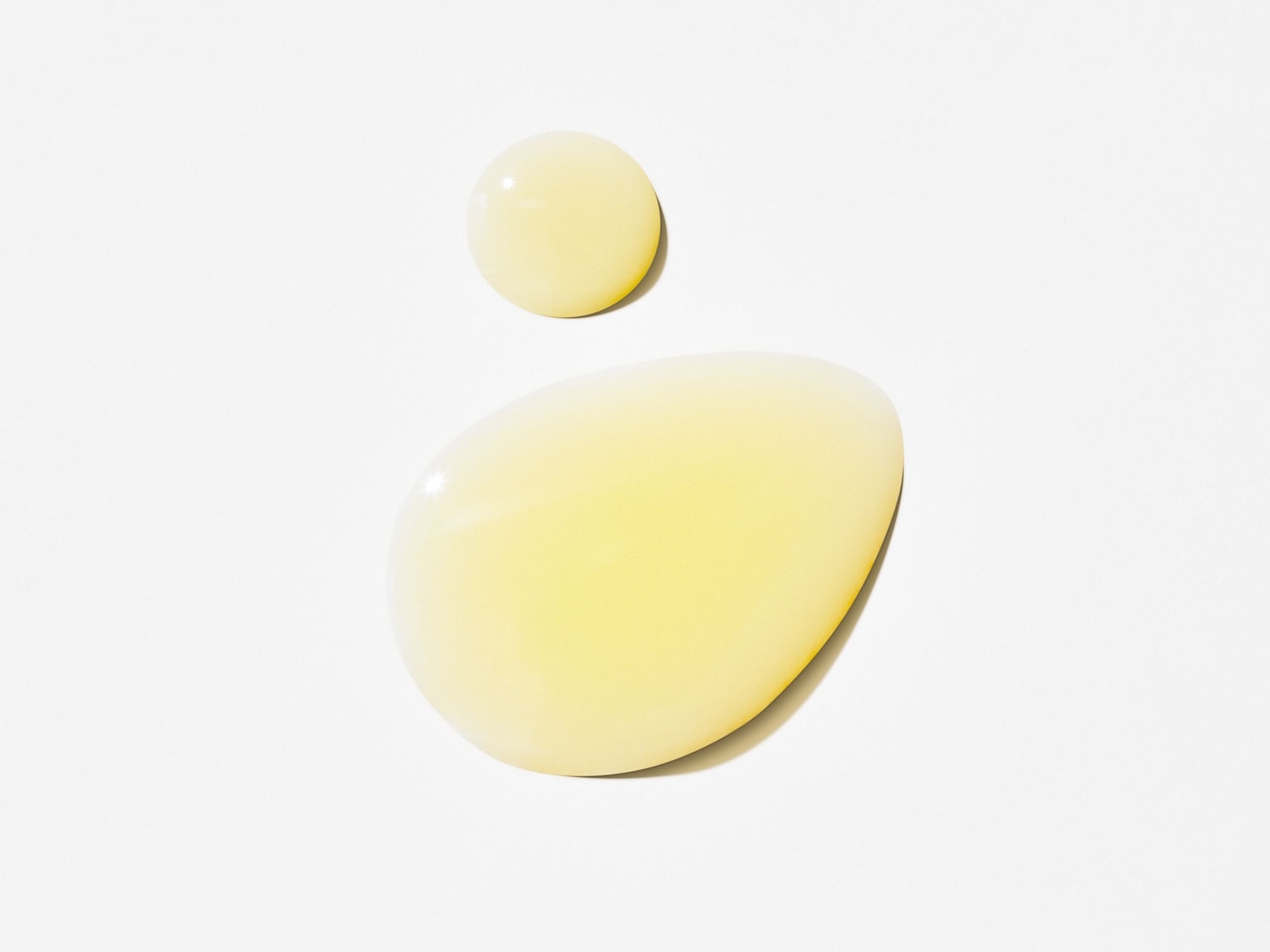 Due gocce di siero dorato su una superficie bianca, una più grande a forma di goccia e una più piccola rotonda, che mostrano la consistenza liscia e luminosa del prodotto.