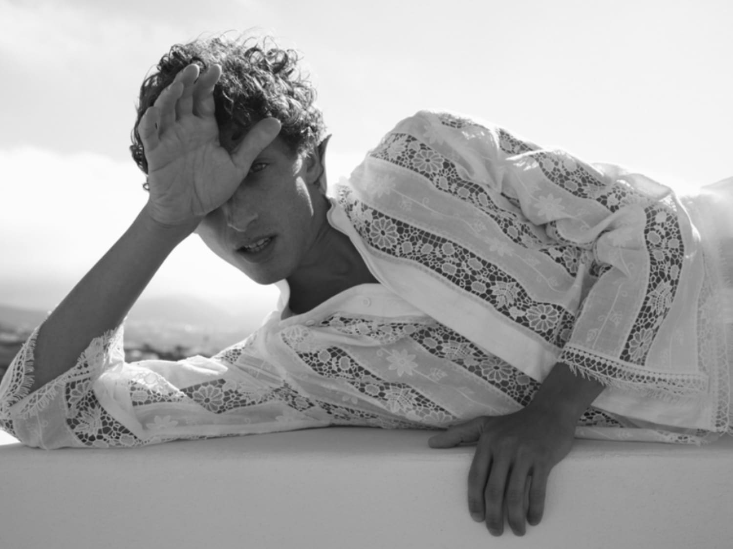 In un elegante scatto in bianco e nero, un uomo con una camicia bianca ricamata si rilassa, con una mano sulla fronte, creando un'atmosfera di sofisticata eleganza.