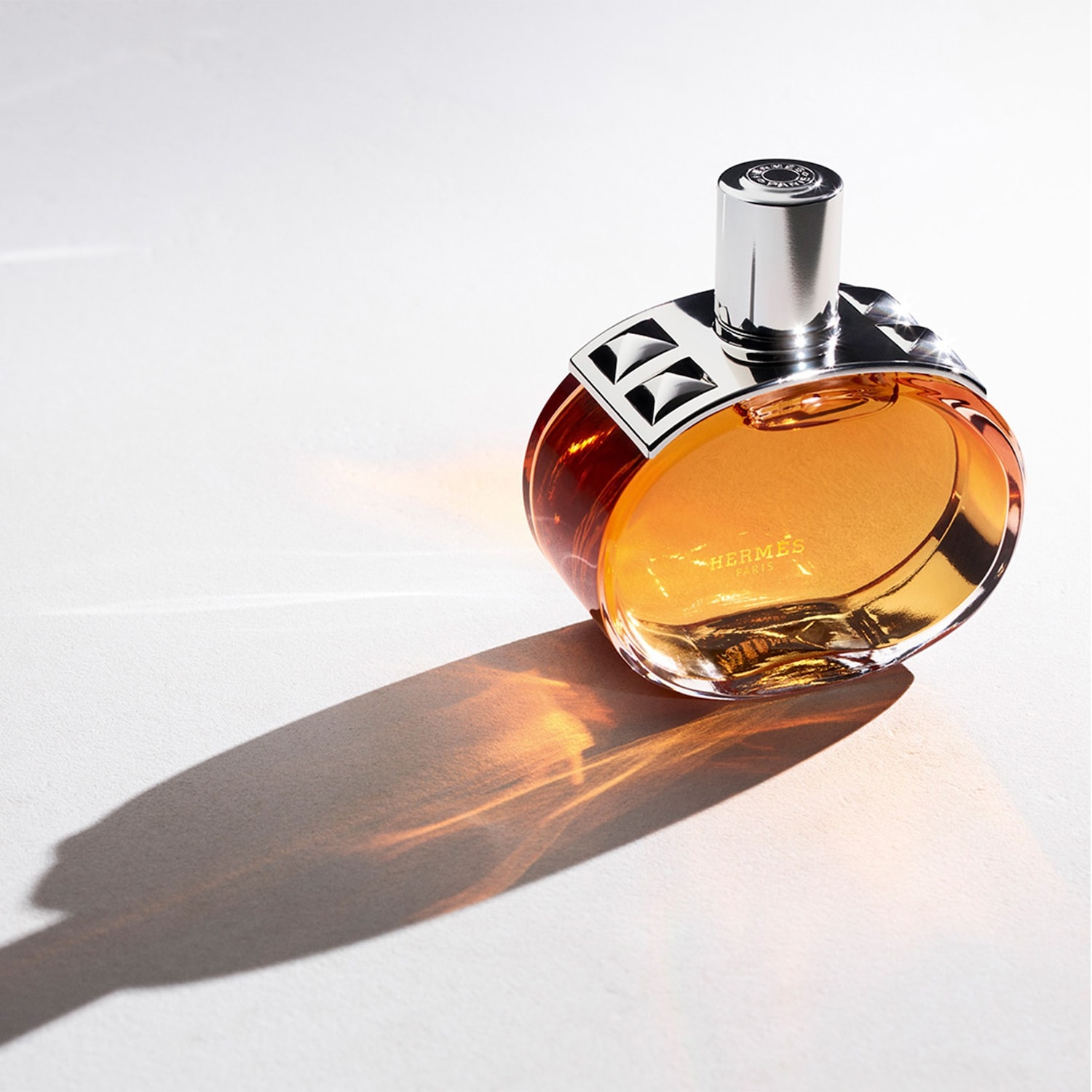 Le parfum Hermès, dans un flacon rond en verre ambré avec un bouchon argenté et un collier clouté, projette une longue ombre sur une surface blanche.