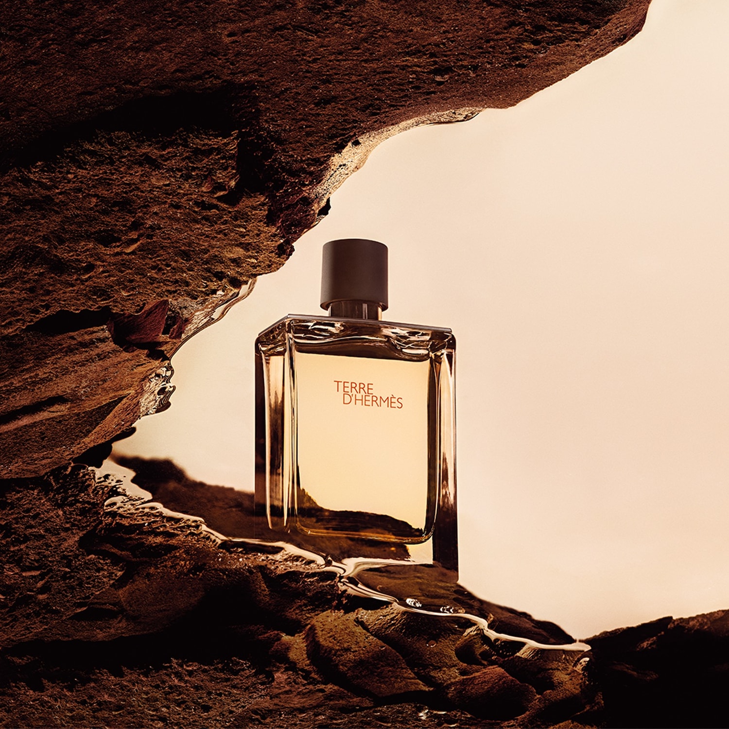 Een heldere glazen fles Terre d'Hermès parfum staat op een rotsachtige ondergrond, met de merknaam in oranje letters en een bruine dop.