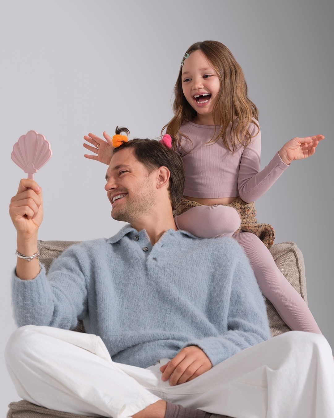 Otec sedí na stoličke s dievčatkom na pleciach, obaja sa usmievajú a majú vo vlasoch farebné gumičky, otec sa pozerá do ružového zrkadla v tvare mušle.