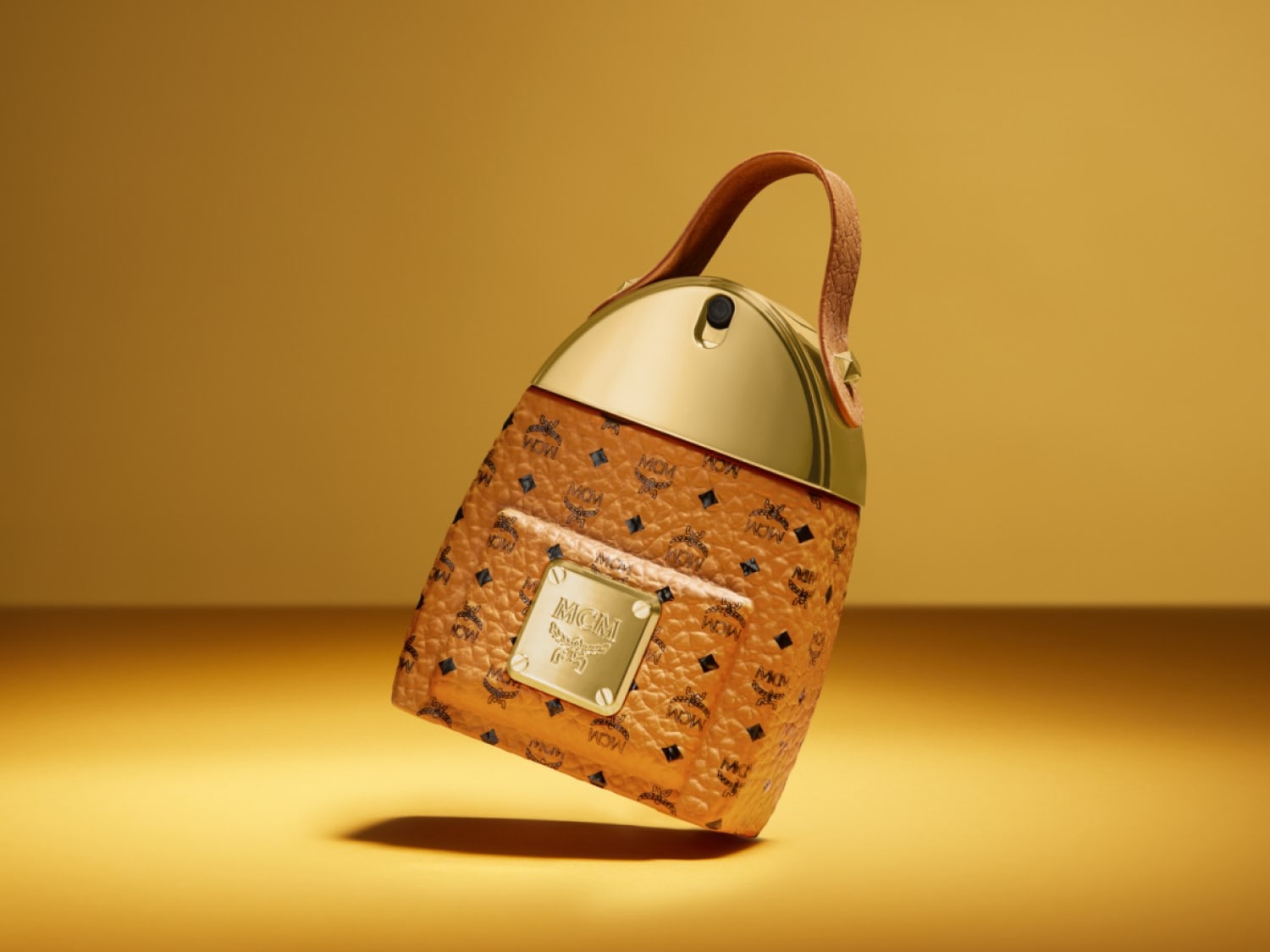 Butelka perfum MCM w kształcie plecaka w kolorze brązowym z logo MCM, złotą górą i brązowym uchwytem na złotym tle.