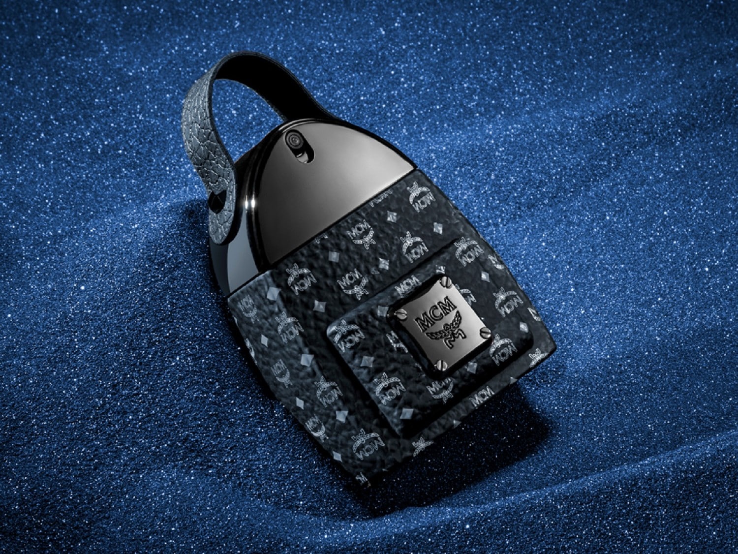 Czarny flakon perfum MCM w kształcie plecaka z srebrnym logo marki na tle błyszczącego, ciemnoniebieskiego brokatu.