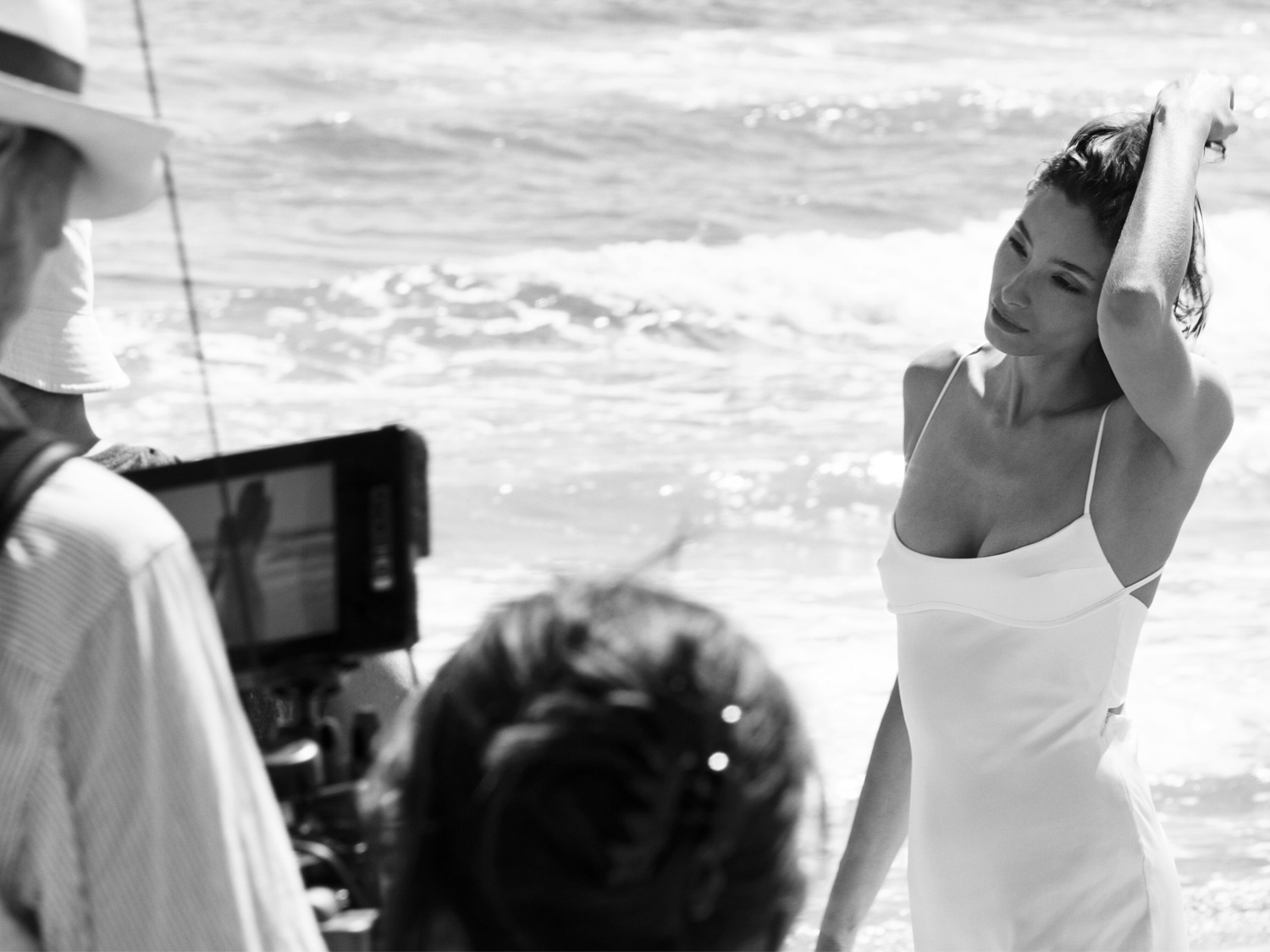 Een zwart-wit foto toont een vrouw in een witte jurk op het strand, gefilmd door een cameraploeg, waarbij de focus ligt op de natuurlijke schoonheid en de serene omgeving.