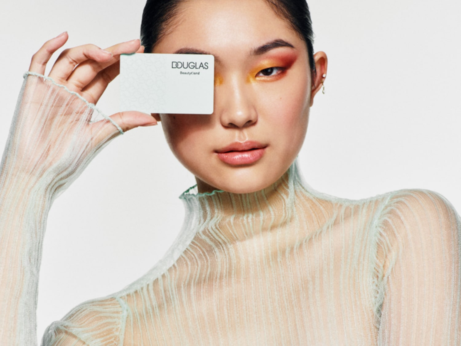 Een vrouw met een doorschijnende top houdt een Douglas Beauty Card voor haar gezicht, met opvallende oranje en rode oogschaduw.