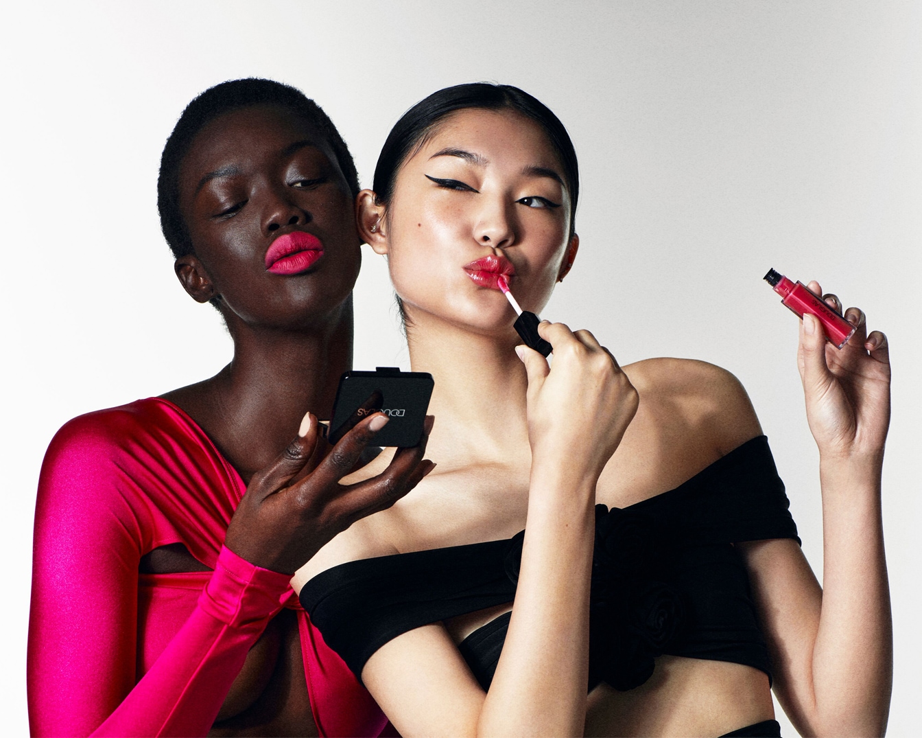 Twee modellen, één met een roze jurk en fuchsia lippenstift die een Douglas compact spiegel vasthoudt, de ander met een zwarte top die rode lipgloss aanbrengt.