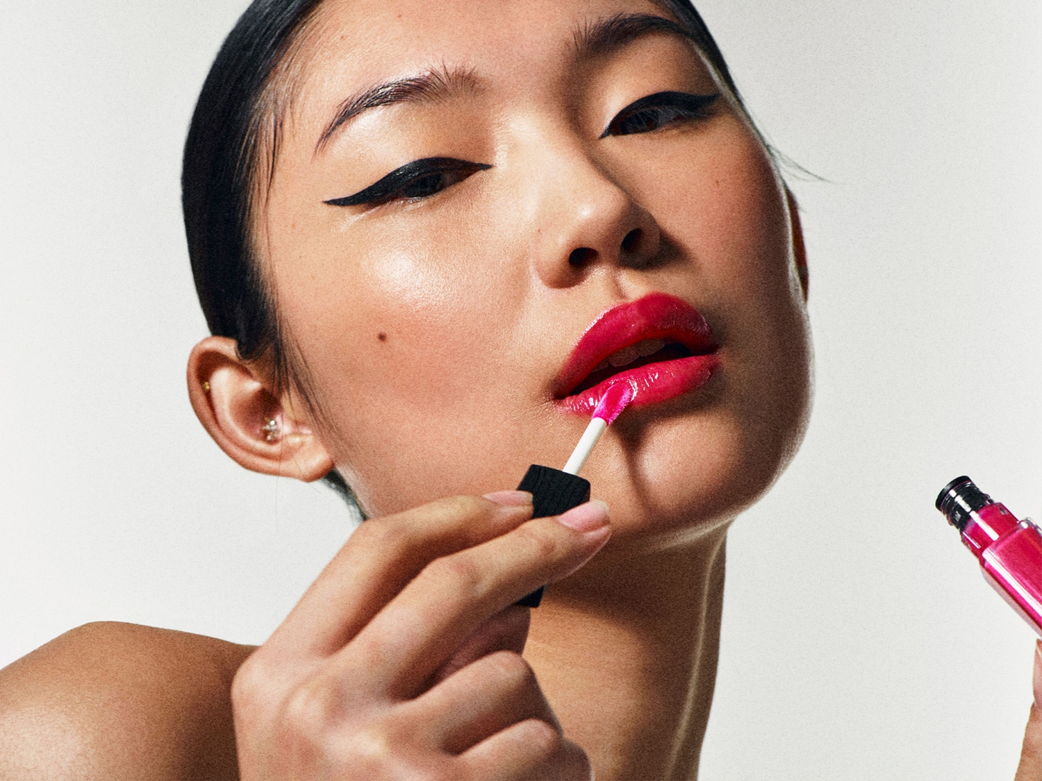 Kobieta z błyszczącymi, różowymi ustami aplikuje błyszczyk aplikatorem z czarną rączką, obok widoczna buteleczka błyszczyku w różowym opakowaniu.