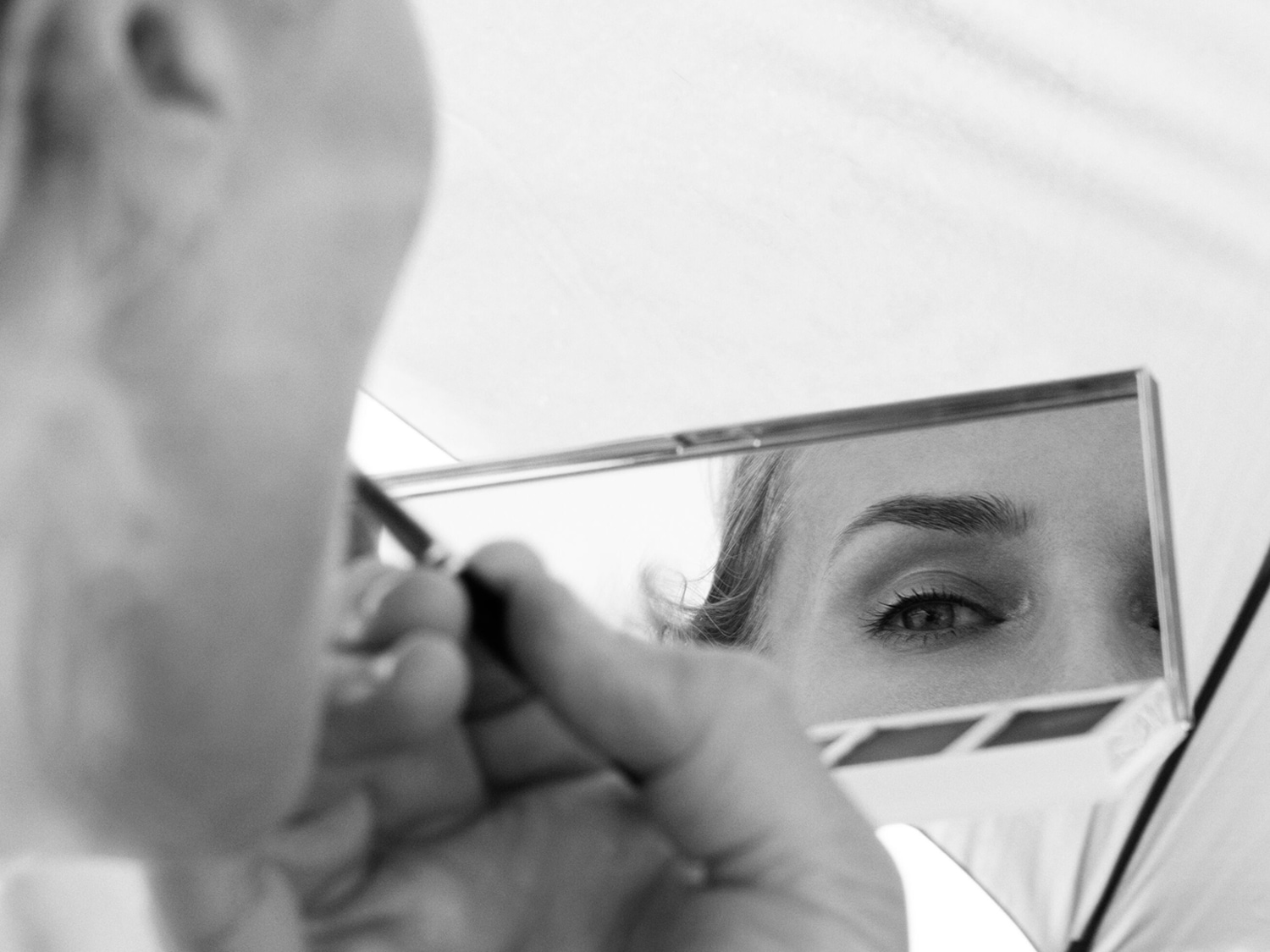 Czarno-białe zdjęcie kobiety, która spogląda w lusterko i podkreśla brwi kredką, skupiając się na precyzyjnym makijażu.