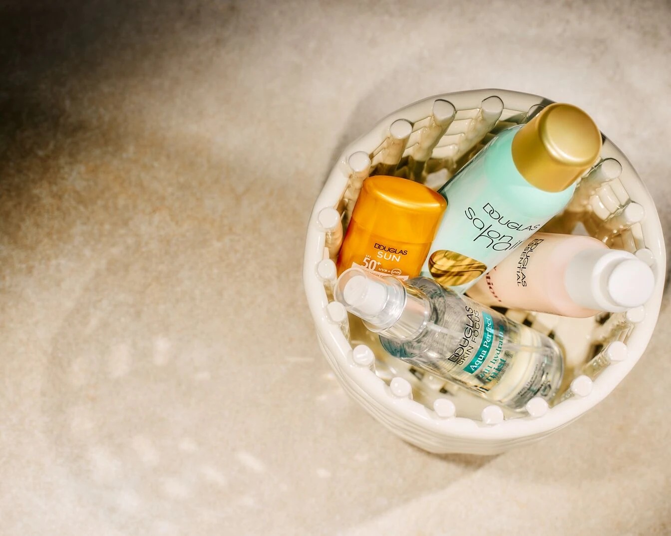 Ein weißer Korb gefüllt mit Douglas Sonnenpflegeprodukten in Orange, Türkis, Rosa und Transparent, die einen umfassenden Schutz und Feuchtigkeitspflege für die Haut bieten.