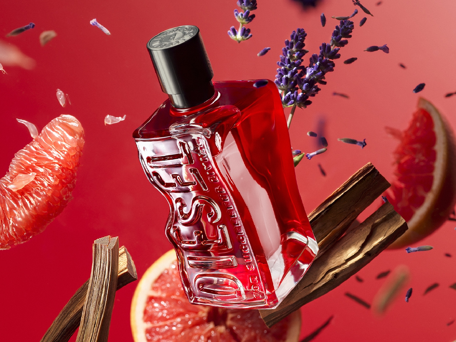 Une bouteille de parfum Diesel rouge et transparente, de forme unique avec le mot 'DIESEL' en relief, flotte au centre d'un fond rouge vif. Autour d'elle flottent des tranches de pamplemousse rose, des bâtons de cannelle, des brins de lavande violette et des gousses de clou de girofle