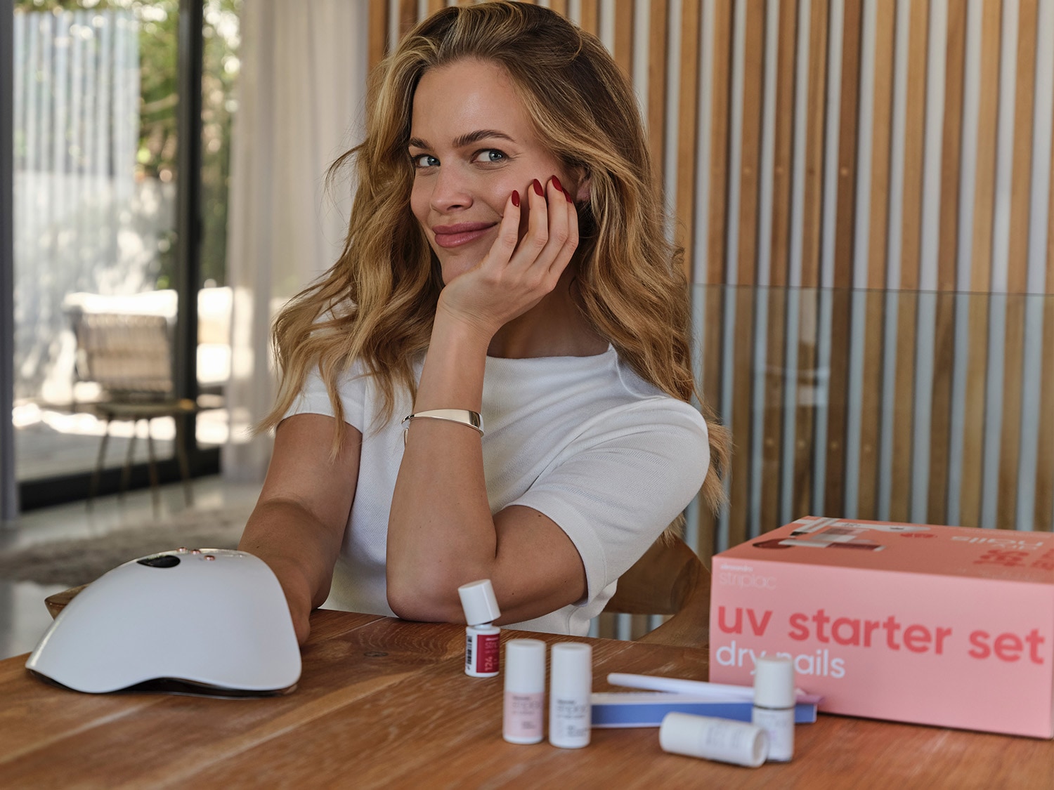 Eine blonde Frau lächelt, während sie ihren Kopf auf ihre Hand stützt, mit einem UV-Nagelset, Nagellackflaschen und einer Lampe auf dem Tisch.