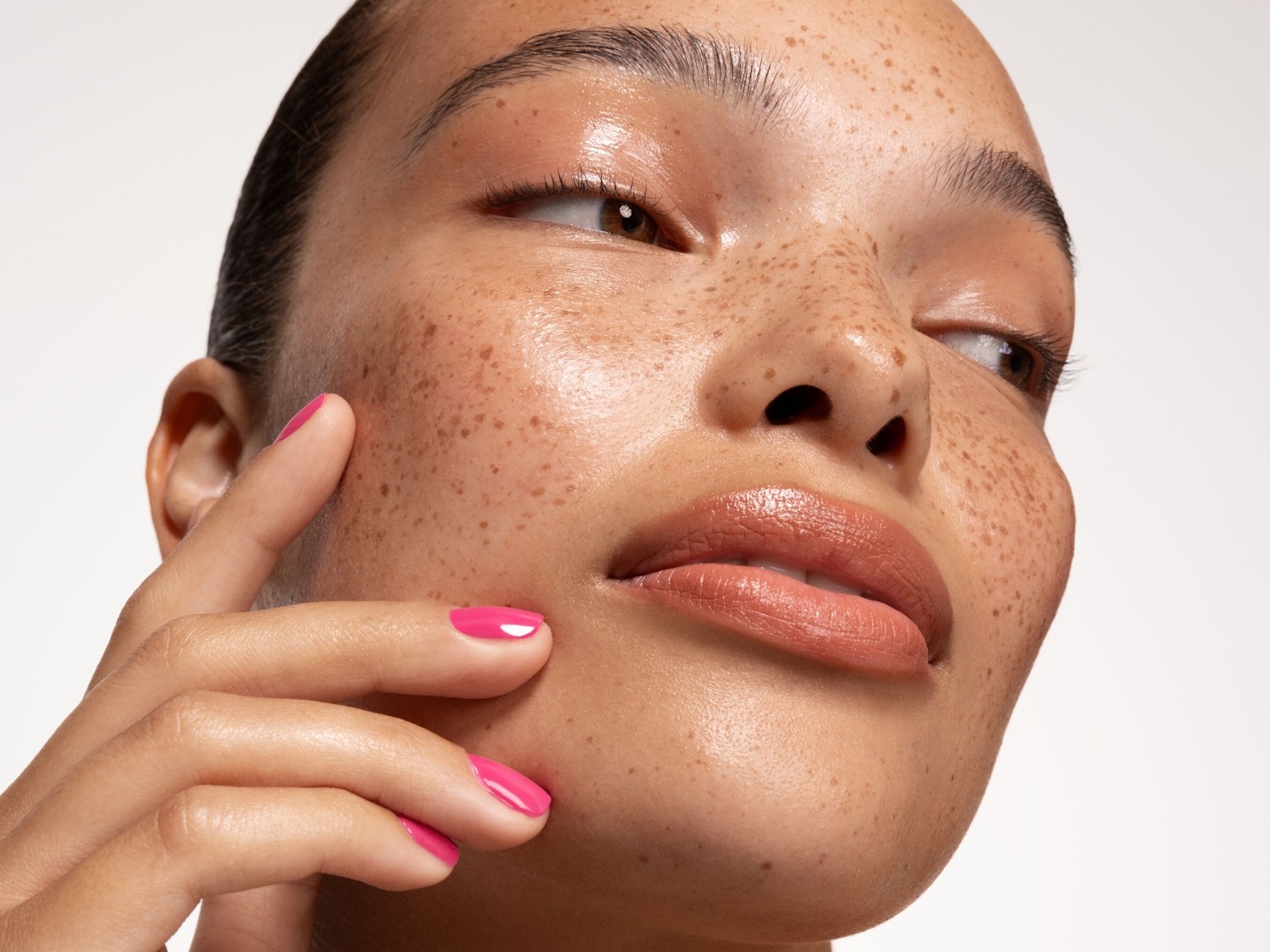 Primer plano del rostro de una mujer con pecas, piel brillante y labios color melocotón, tocándose la mejilla con una mano de uñas rosas.