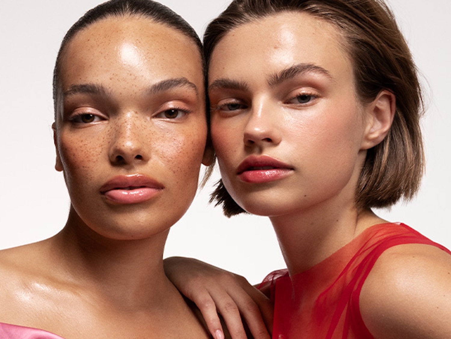 Dos modelos con maquillaje natural resaltan sus rasgos; una luce un vestido rosa y la otra uno rojo, ambas con labios brillantes y piel luminosa.