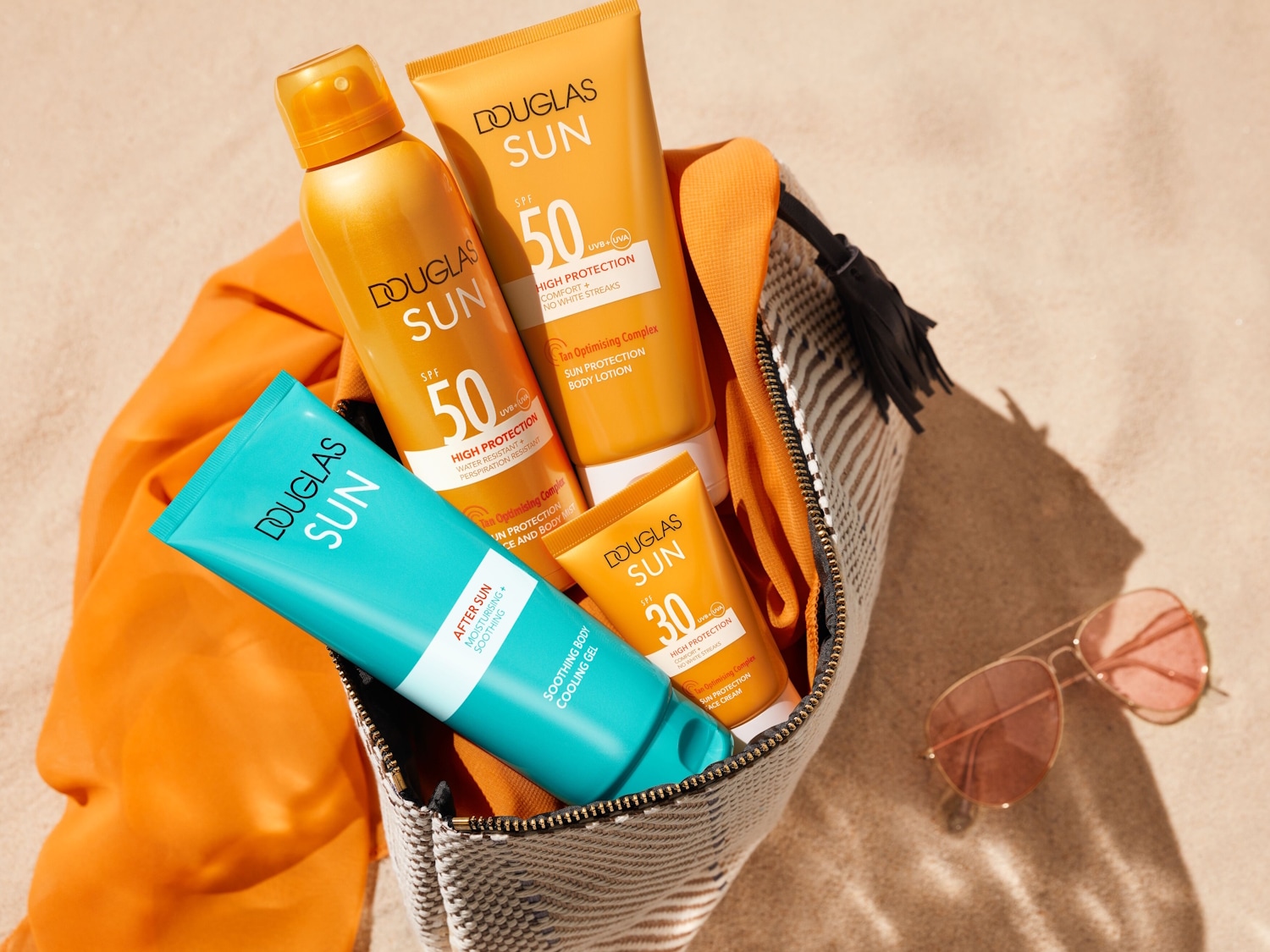 Paplūdimio krepšyje matomi „Douglas Sun“ saulės priežiūros produktai: purškiklis SPF 50, losjonas SPF 50, kremas SPF 30 ir raminantis gelis po saulės vonių.