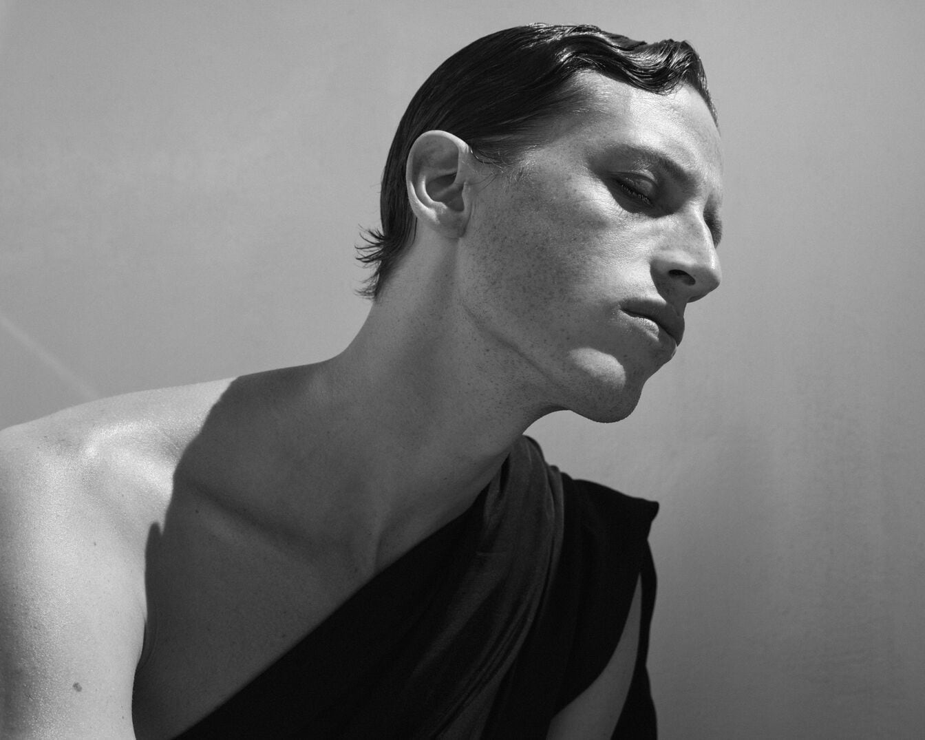 Juodai balta nuotrauka, kurioje matomas vyras, atsukęs galvą į šoną, su šukuosena ir tamsiu audeklu ant peties.