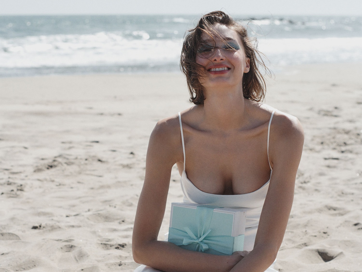 Eine lächelnde Frau im weißen Kleid sitzt am Strand und hält eine hellblaue Geschenkbox mit Schleife vor dem Hintergrund von Sand und Meer.
