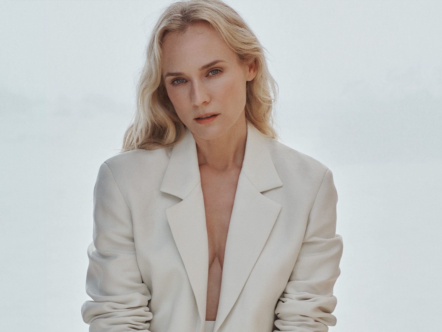 Blonde Frau mit blauen Augen, die einen weißen Blazer ohne Hemd darunter trägt und direkt in die Kamera schaut.