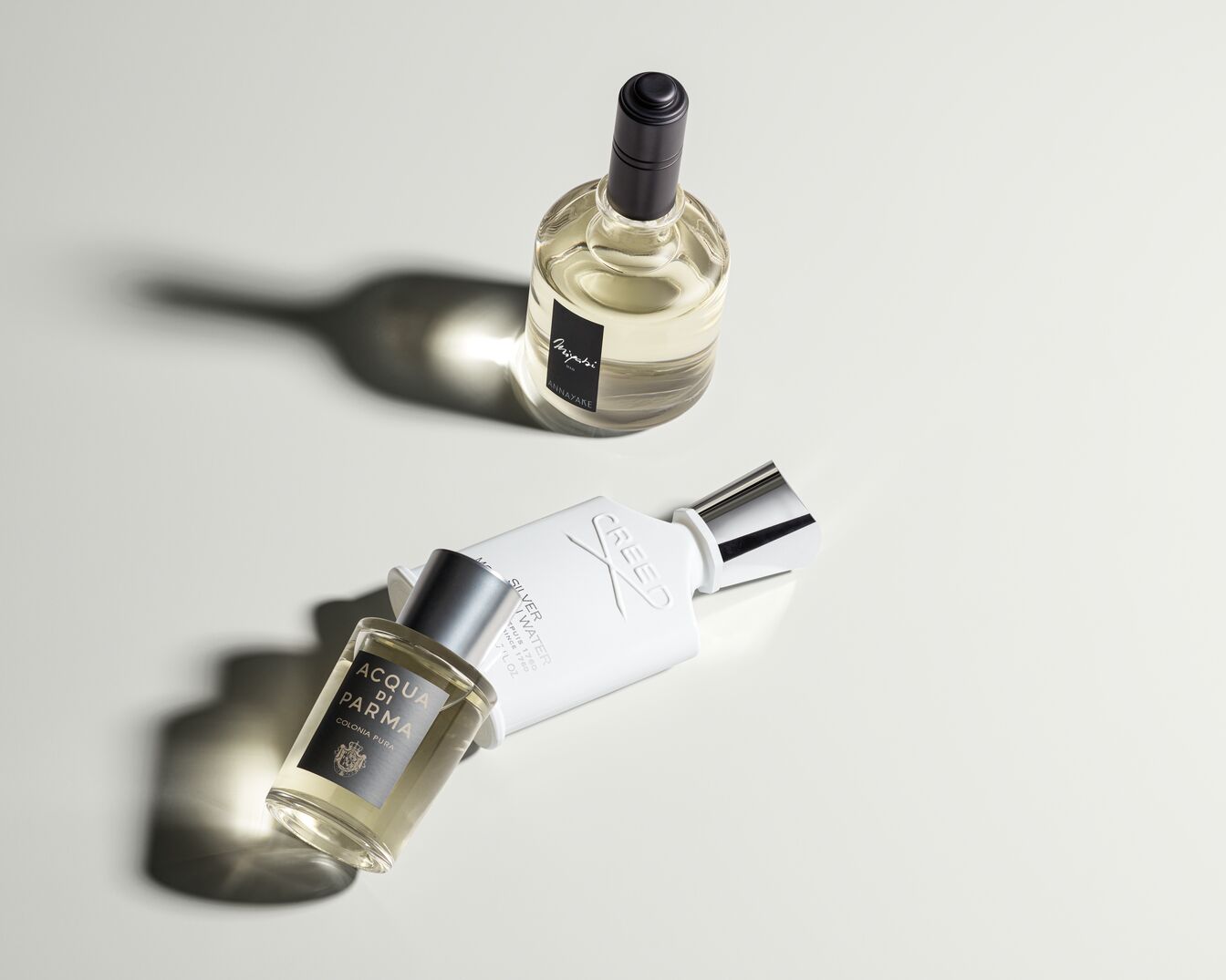 Nuotraukoje matomi trys kvepalų buteliukai: Miyabi Annayake, Acqua di Parma Colonia Pura ir Creed Silver Water, išdėstyti ant balto paviršiaus.