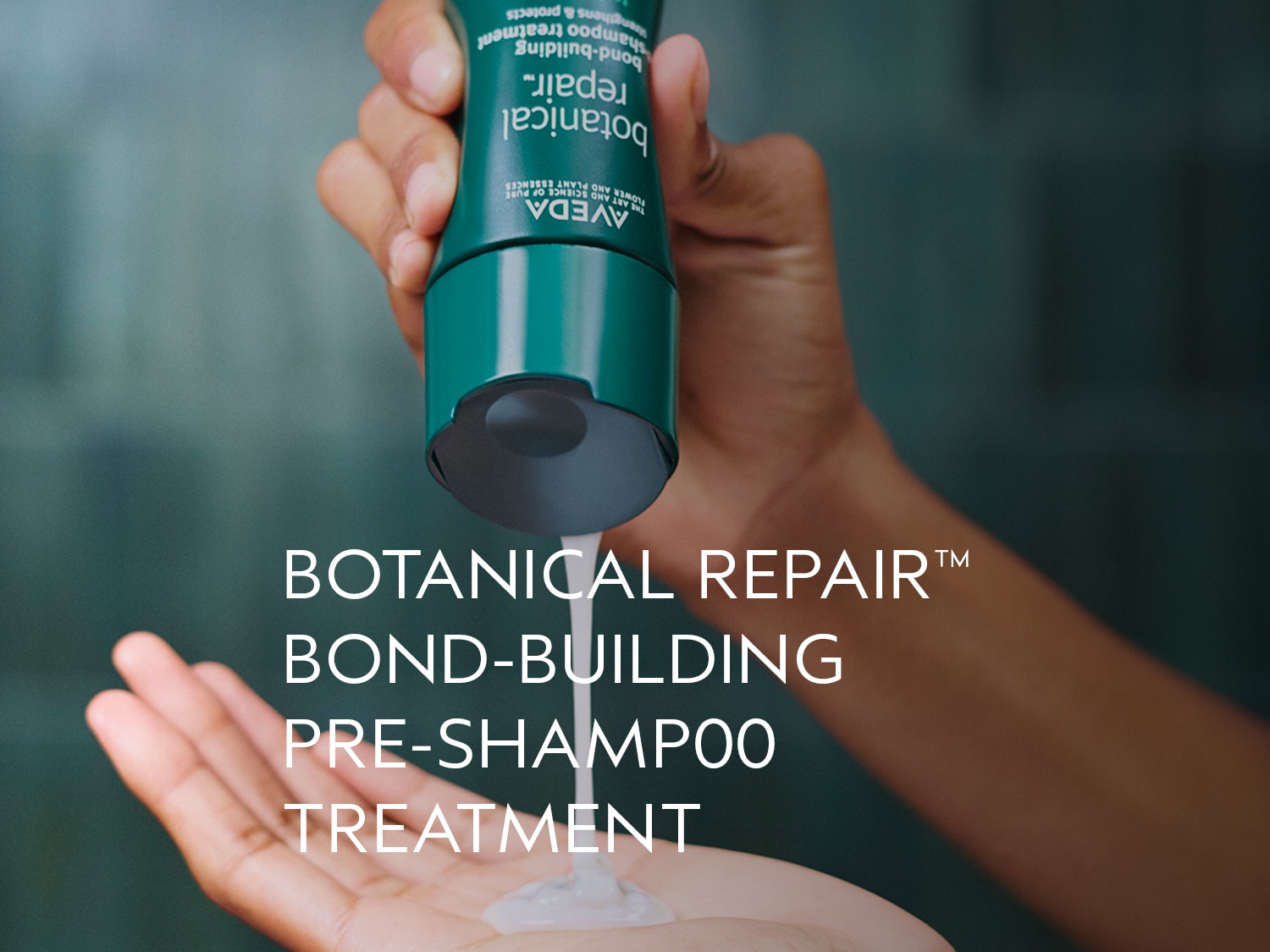 Een hand houdt een groene tube Aveda Botanical Repair Pre-Shampoo Treatment vast, terwijl een witte crème in de handpalm wordt gegoten.