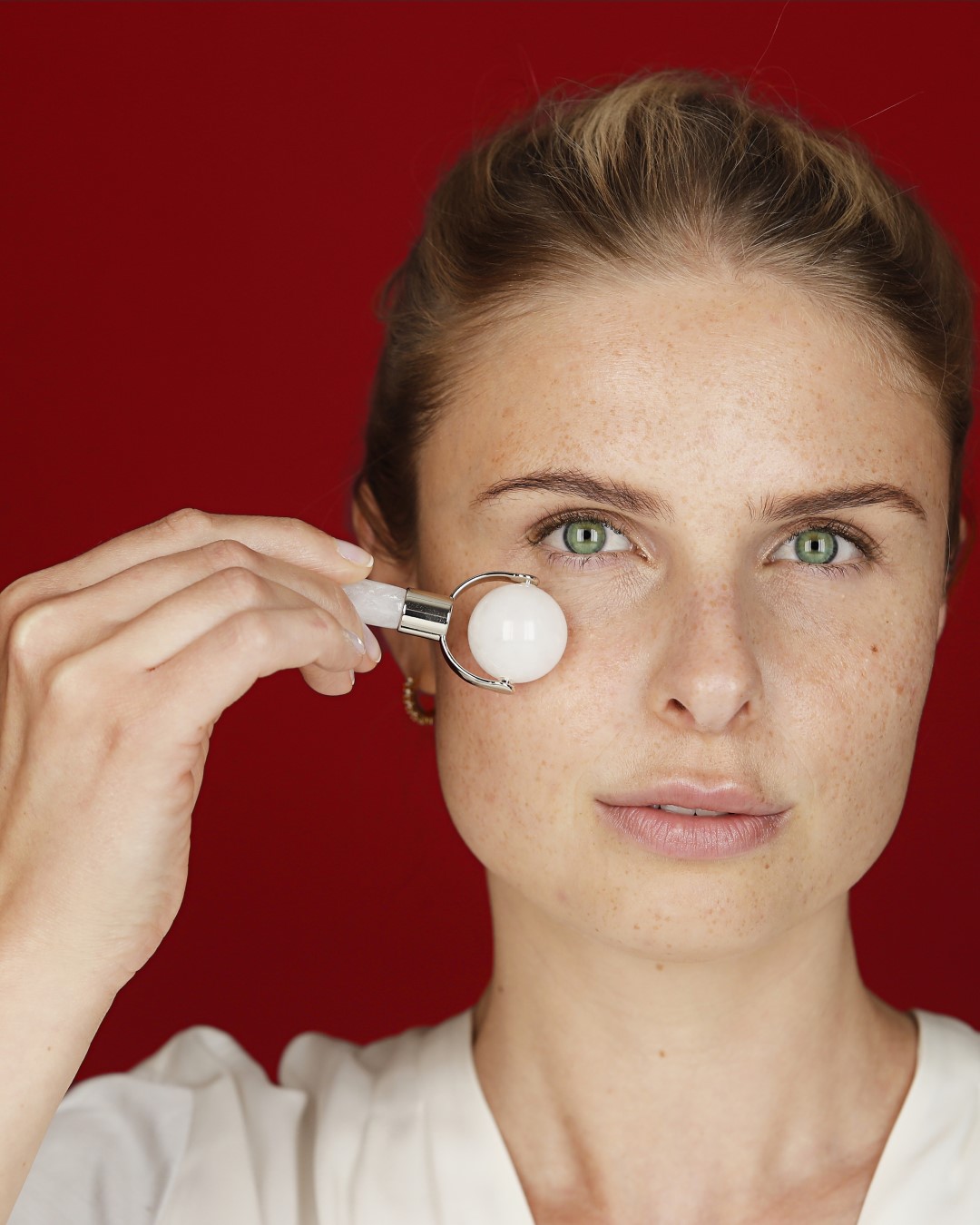 Eine Frau mit hellen Haaren und Sommersprossen benutzt einen weißen Jade-Gesichtsroller auf ihrer Wange vor einem roten Hintergrund.